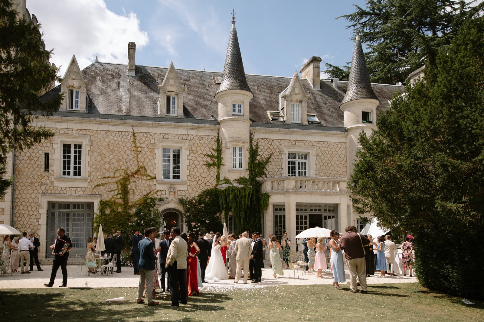 A group of people, many dressed formally, gather outside a large, stone mansion with turrets and ivy-covered walls on a sunny day. Chateau de la Couronne wedding.