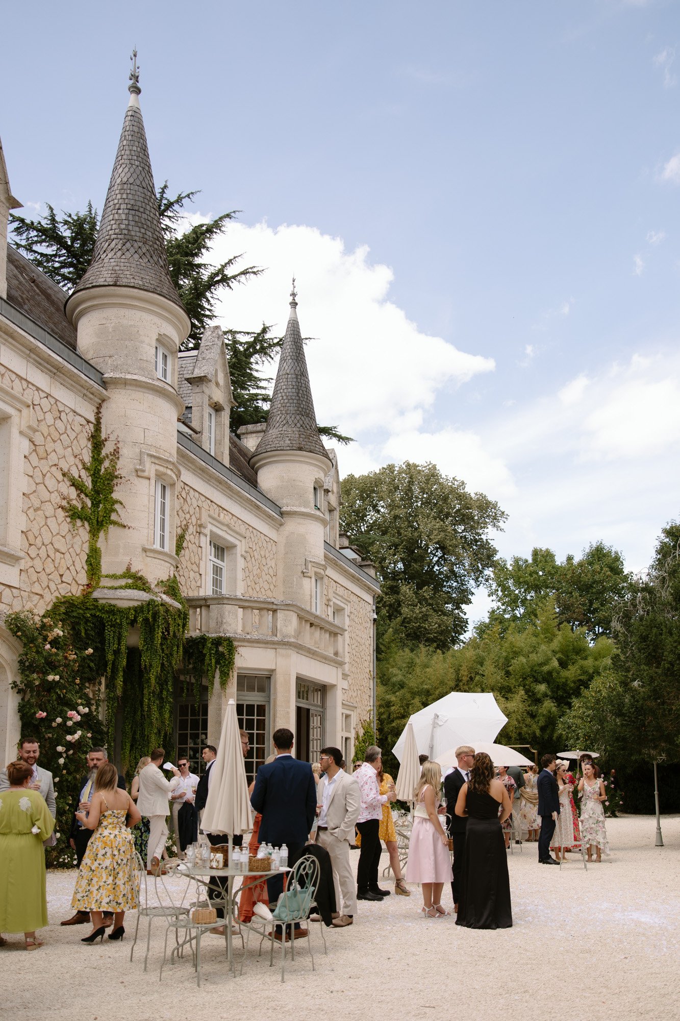 A group of people in formal attire gather and converse outside a stone château with turrets on a sunny day. Chateau de la Couronne wedding.