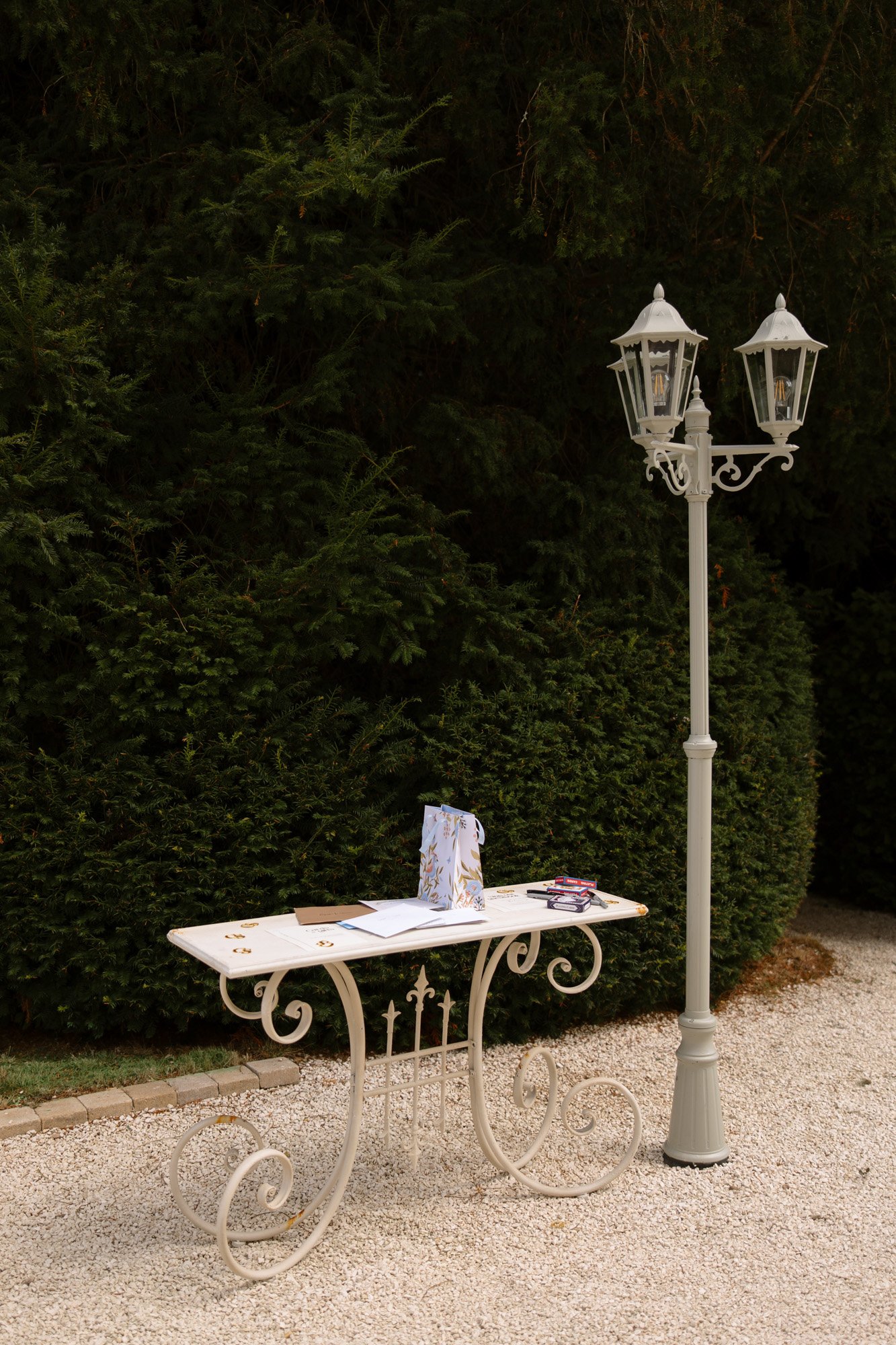 A white metal table with papers and a gift bag stands on gravel next to a vintage street lamp, with dense green bushes in the background.