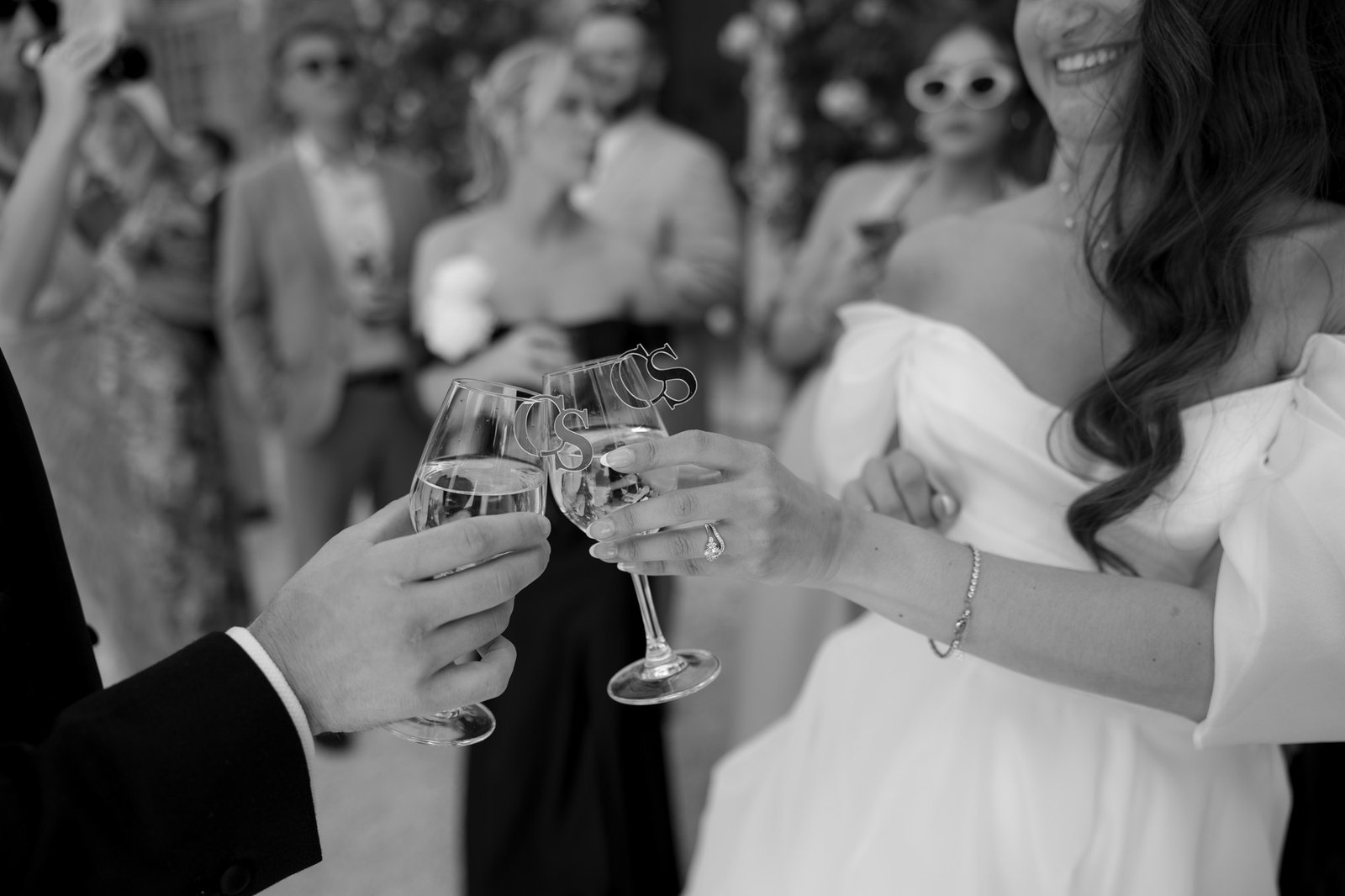 A bride and another person clink champagne glasses at a celebration, with guests in formal attire blurred in the background. Chateau de la Couronne wedding.