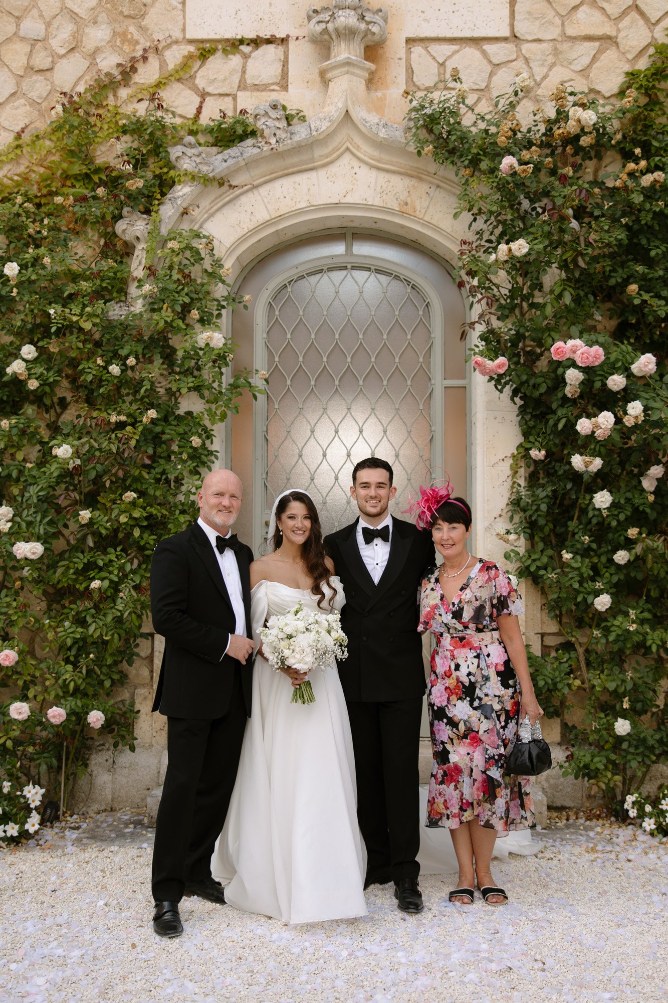 Four people pose in front of a stone building with roses. Two men wear black tuxedos, while two women wear dresses; one is in a white bridal gown holding a bouquet.