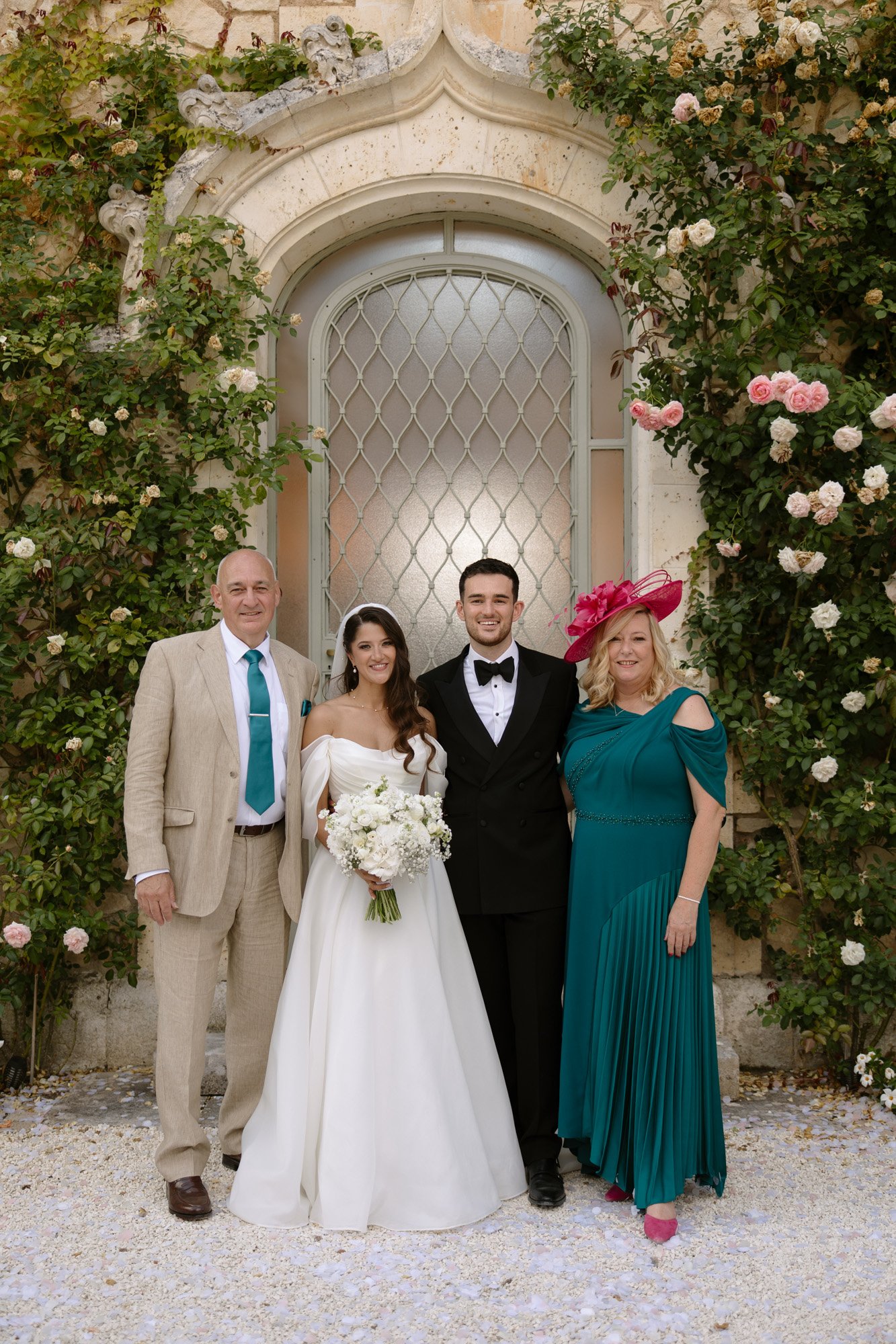 Four people pose in front of a stone building with climbing roses; the bride and groom stand in the middle, flanked by two adults dressed formally.