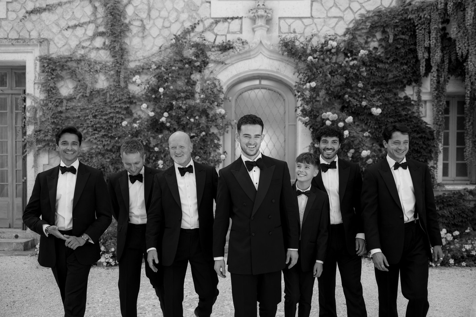 Seven men dressed in tuxedos stand and walk together outside a stone building covered in ivy, posing for a formal group photo. Chateau de la Couronne wedding.