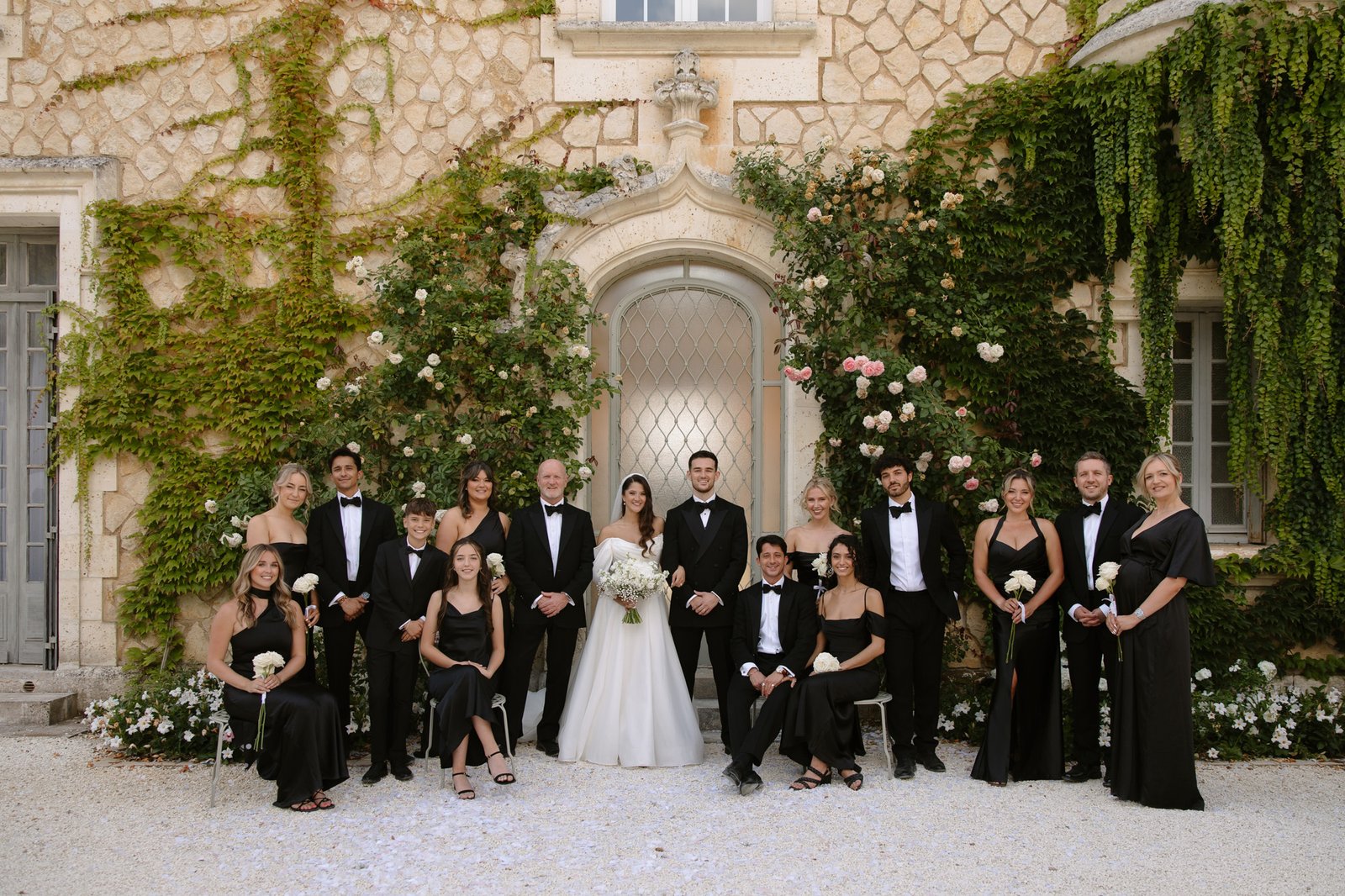 A bride and groom stand with their wedding party dressed in black attire, posing for a formal group photo outside an ivy-covered stone building.