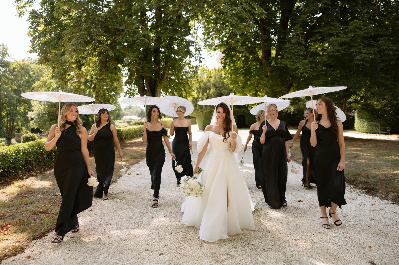 A bride in a white gown walks with bridesmaids in black dresses and white parasols along a tree-lined path on a sunny day. Chateau de la Couronne wedding.
