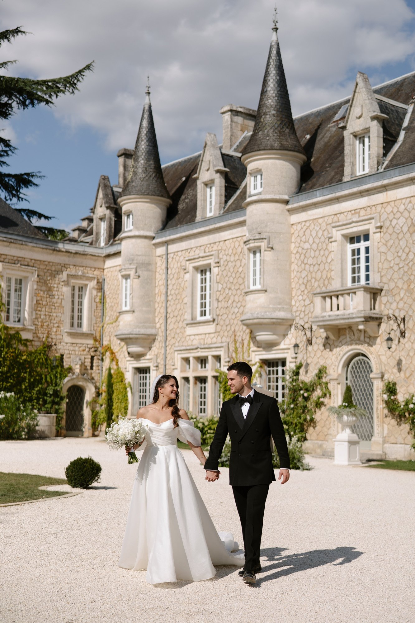 A bride and groom in formal attire walk hand in hand outside a historic stone chateau with turrets on a sunny day. Chateau de la Couronne wedding.