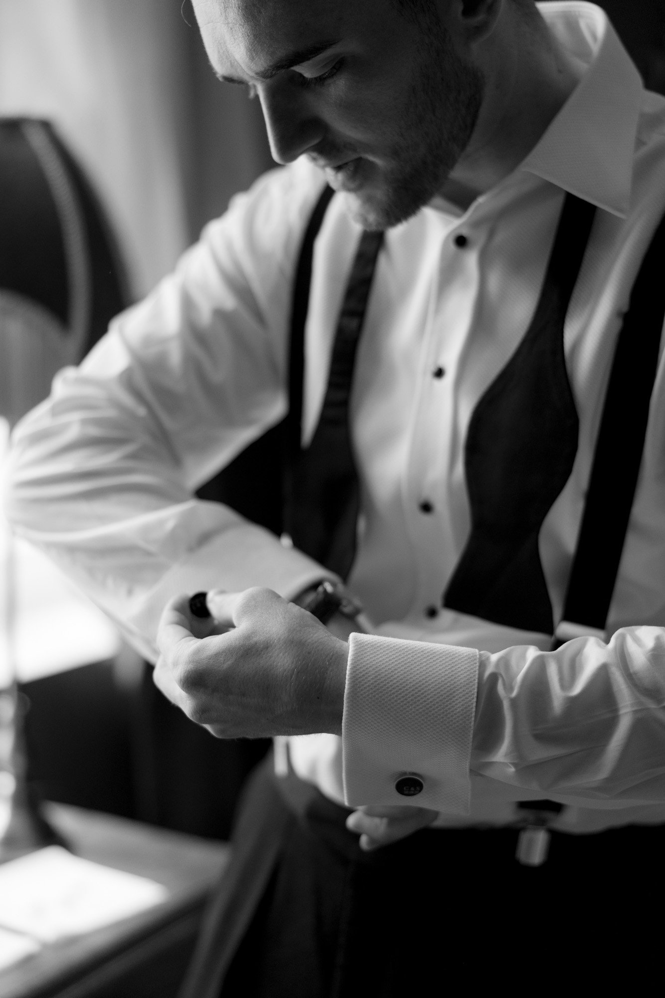 A man in formal attire adjusts his cufflink while wearing an unfastened bow tie in a softly lit room.