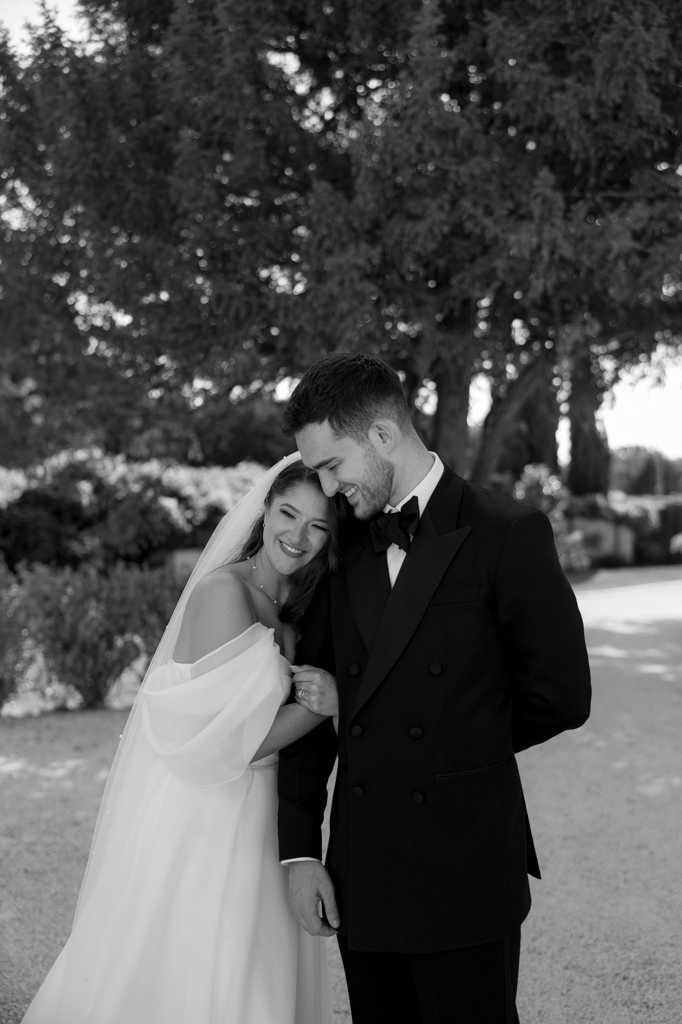 A bride in a white dress and veil smiles, leaning on a groom in a black suit, standing outdoors with trees and bushes in the background. Chateau de la Couronne wedding.