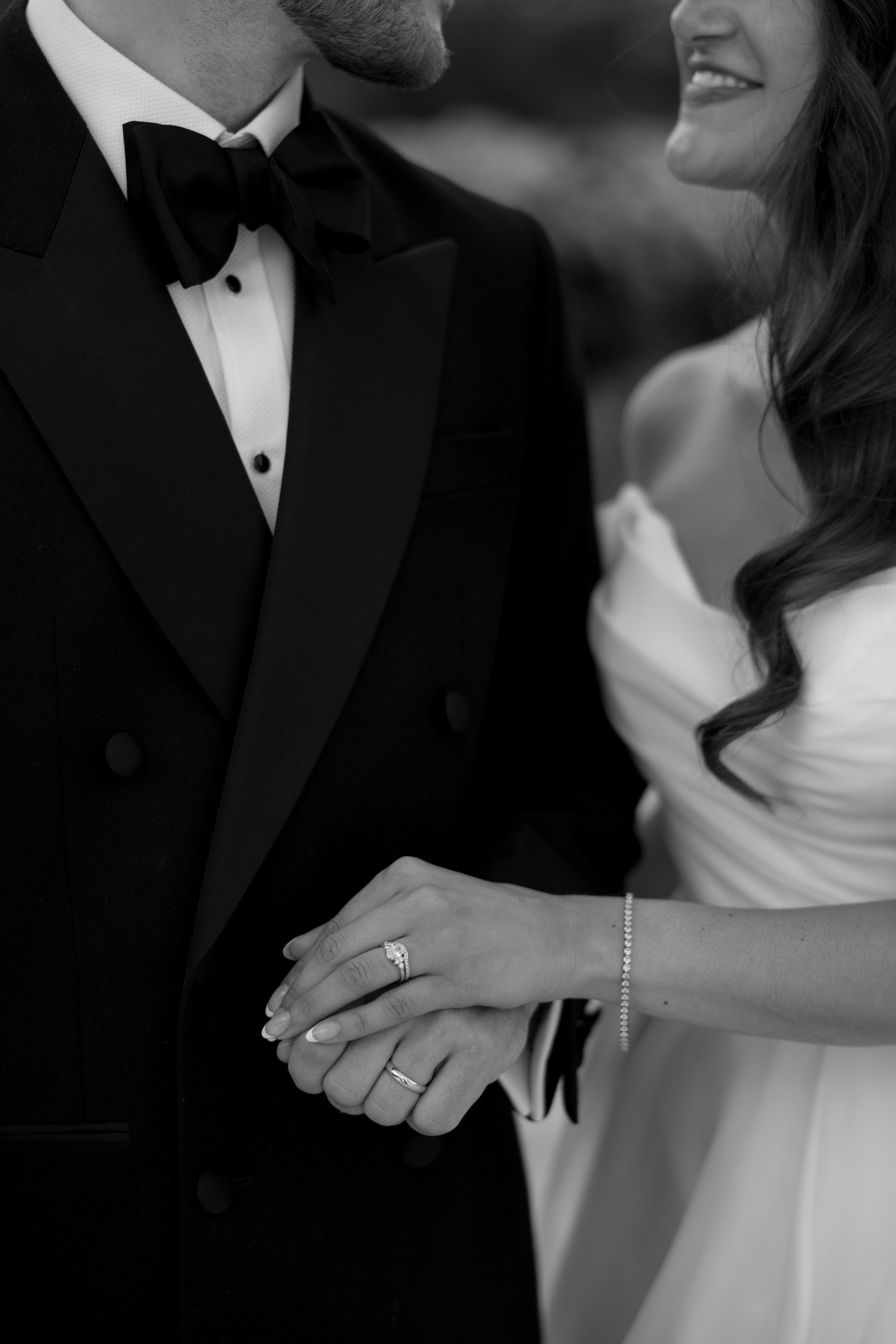 A couple dressed in formal attire holds hands, showing wedding rings. The image is in black and white and their faces are partially out of frame. Chateau de la Couronne wedding.