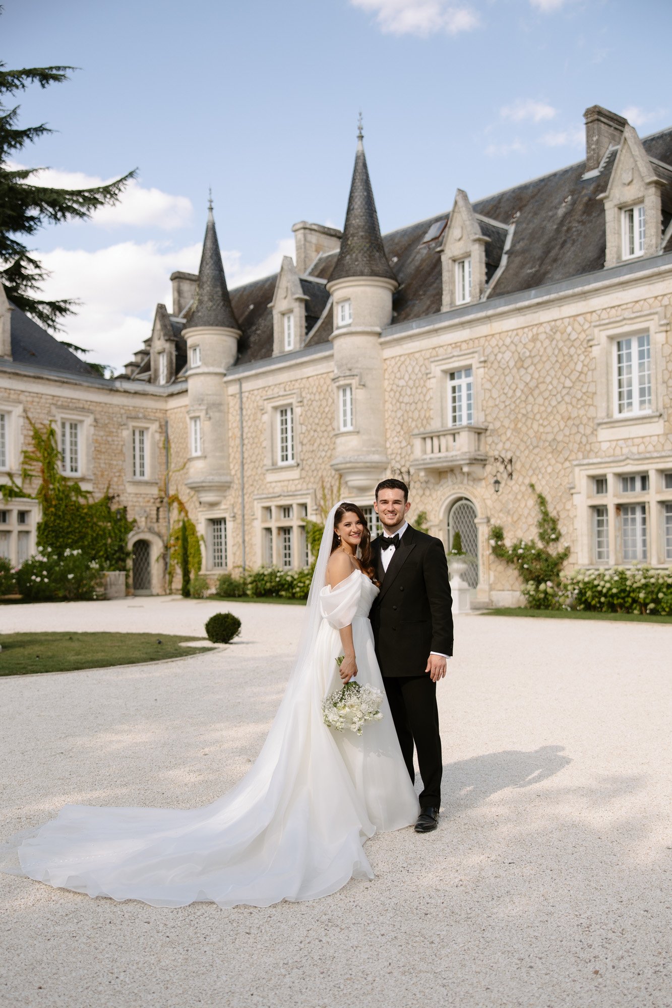 A bride and groom pose together in wedding attire outside a large stone chateau with tall peaked roofs on a sunny day. Chateau de la Couronne wedding.