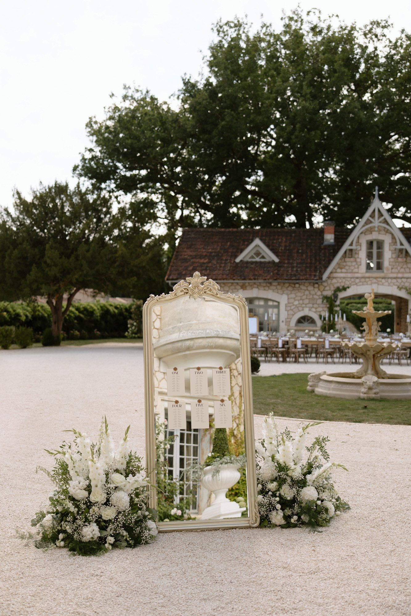 A tall ornate mirror stands outdoors on gravel, framed by white floral arrangements, reflecting a decorative structure; a stone building and fountain are in the background. Chateau de la Couronne wedding.