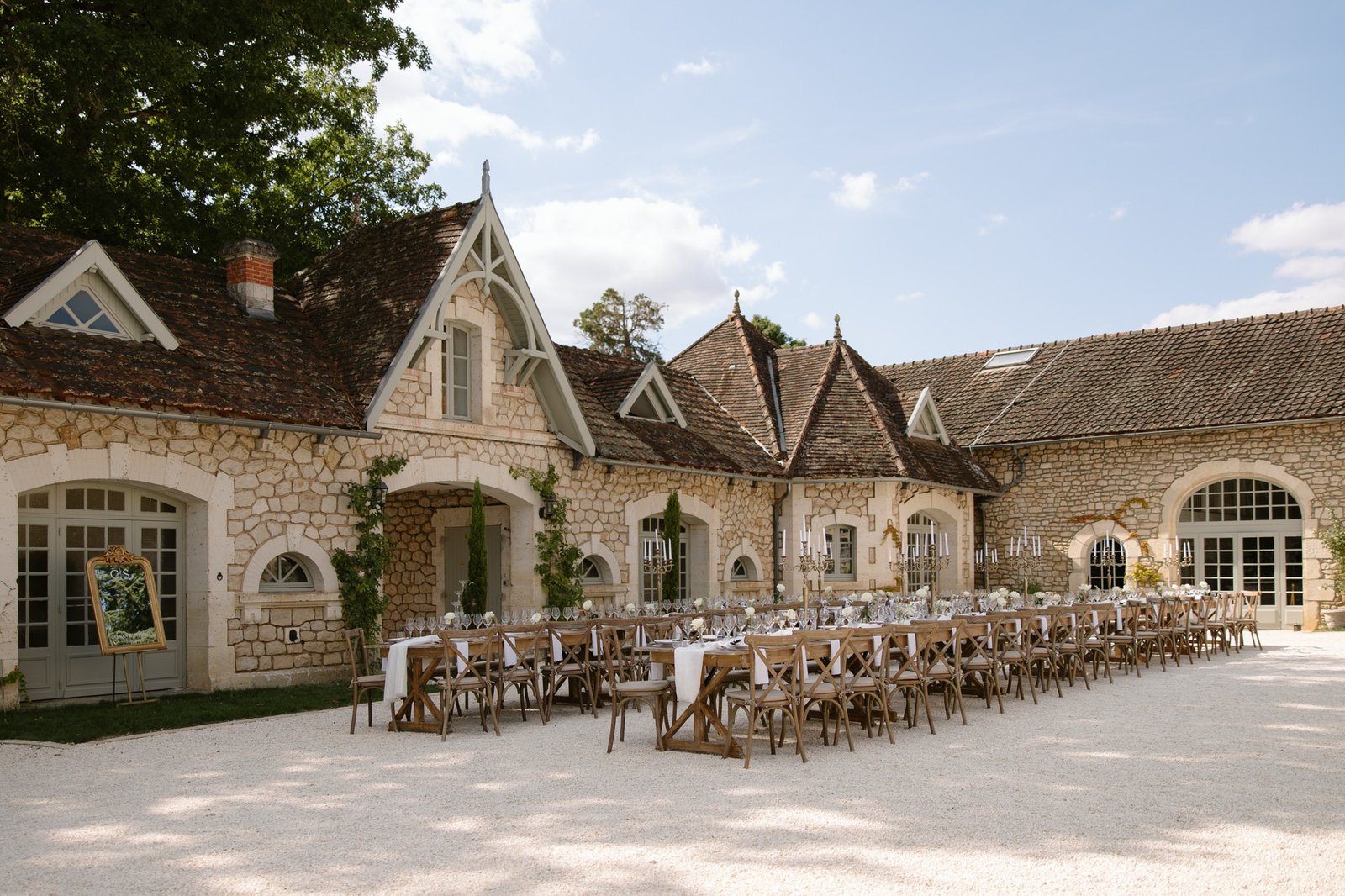 Long banquet tables with wooden chairs are set up in the courtyard of a rustic stone building under a partly cloudy sky. Chateau de la Couronne wedding.