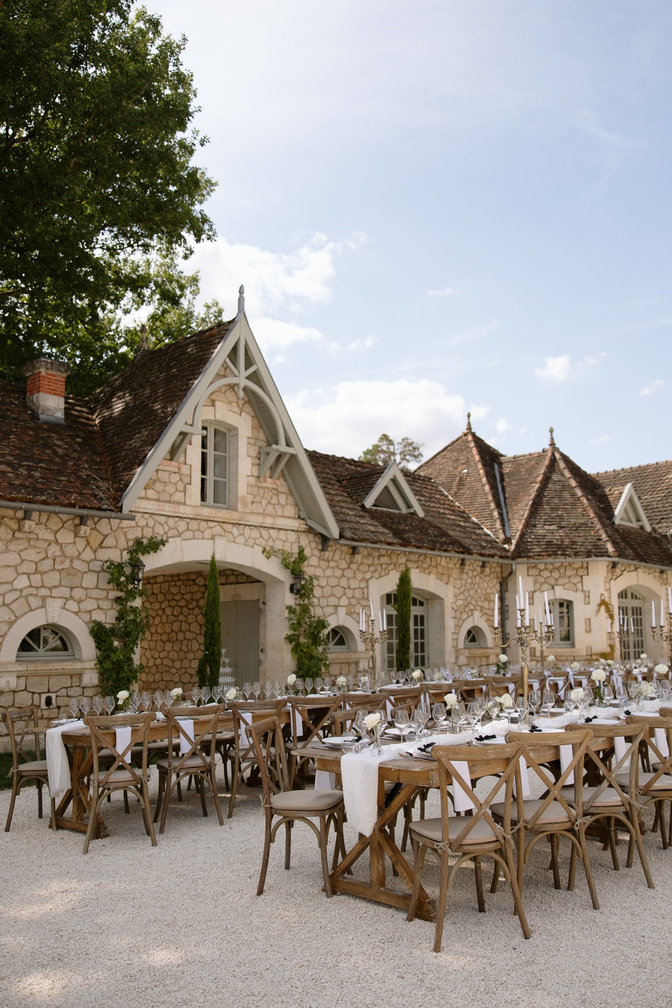 Outdoor dining tables set for an event are arranged in front of a rustic stone building with pitched roofs and greenery. Chateau de la Couronne wedding.
