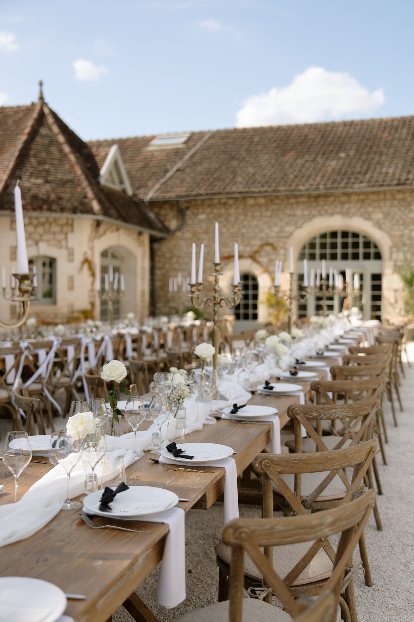 Long wooden tables with white table settings, glassware, and floral centerpieces are arranged outdoors in a courtyard with rustic stone buildings and candelabras. Chateau de la Couronne wedding.
