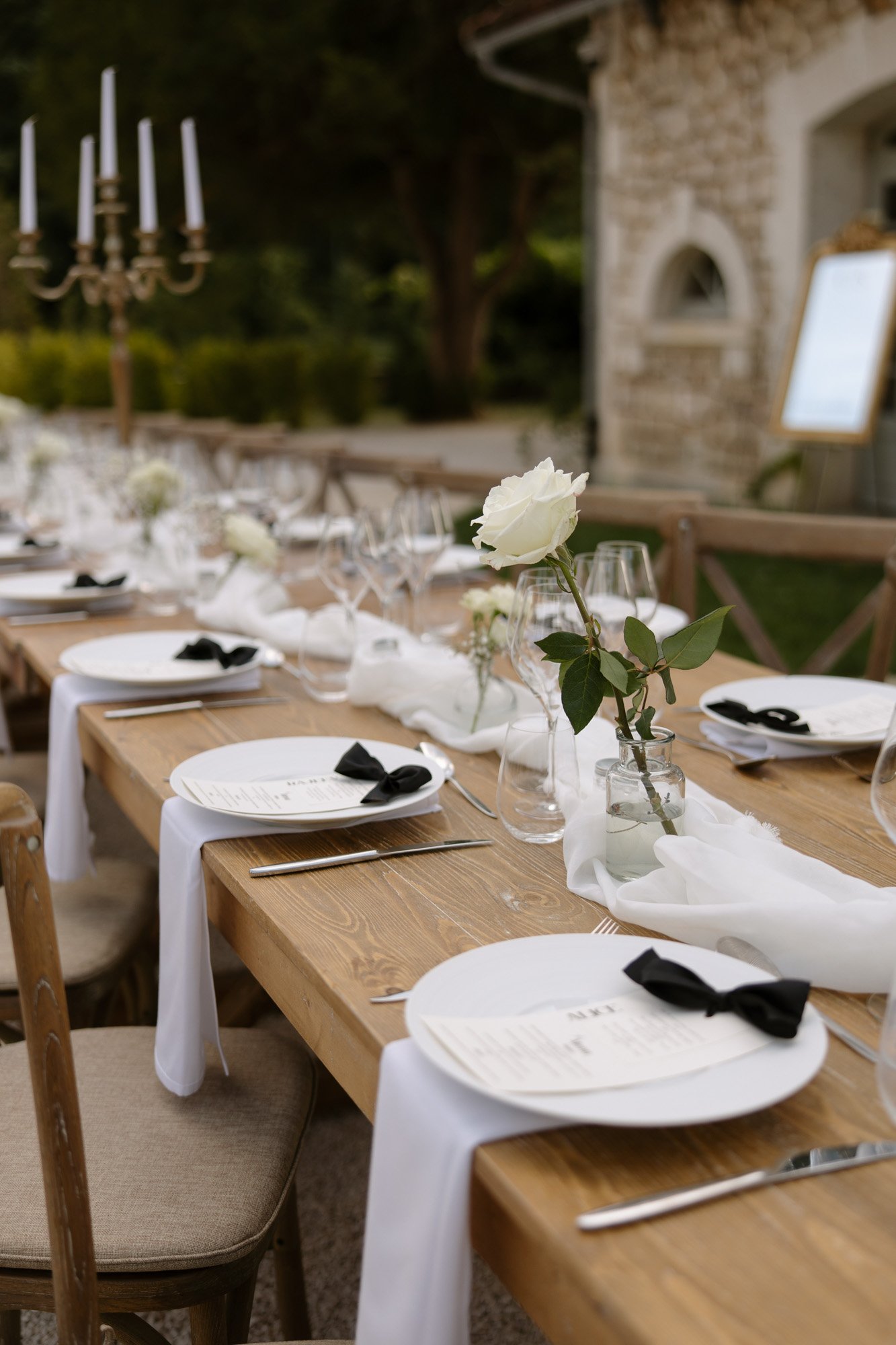 A long wooden table set for an outdoor event with white plates, black napkins, wine glasses, white roses in vases, and white cloth runners.