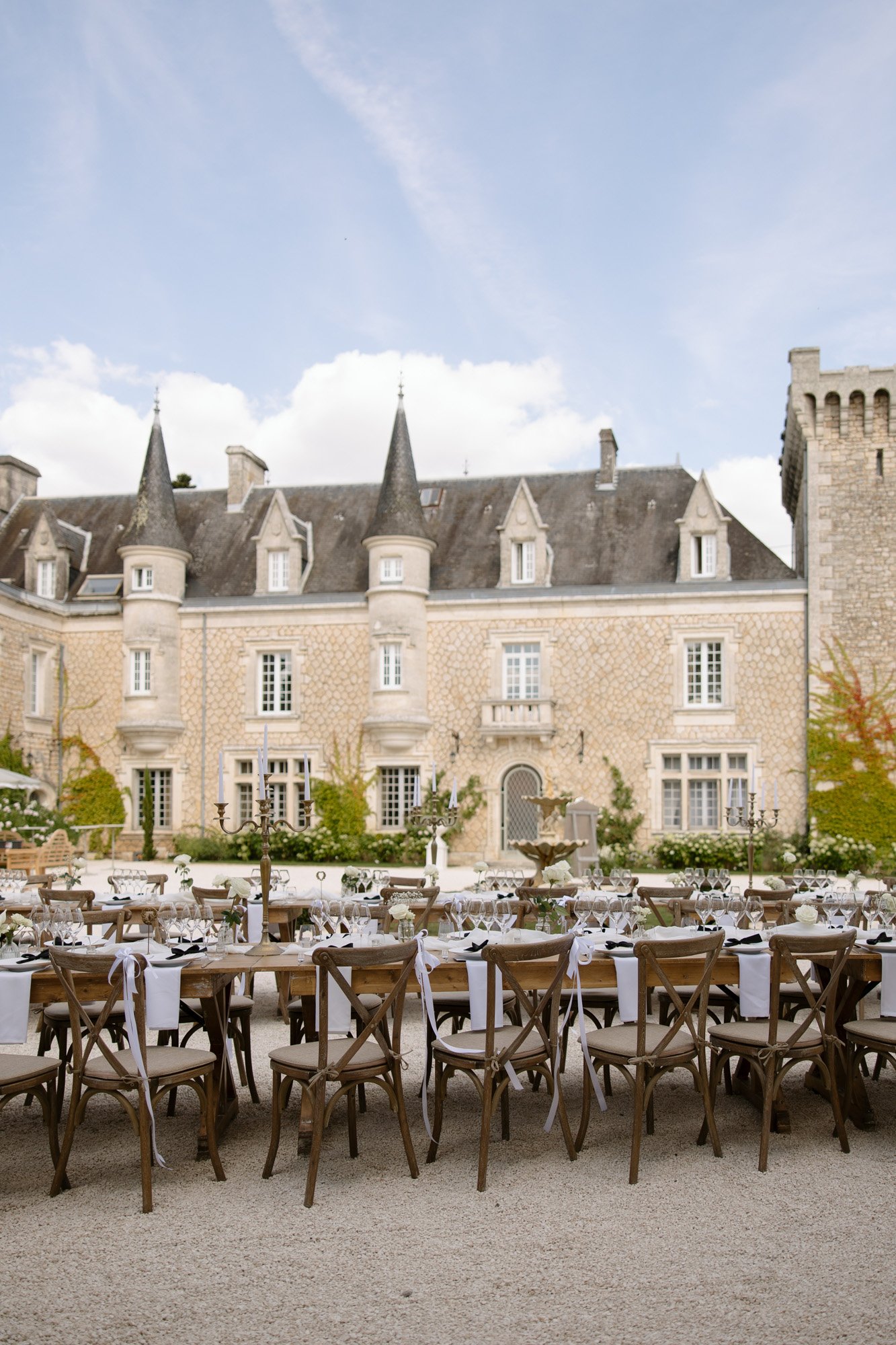 Long banquet tables set for an outdoor event are arranged in front of a historic stone chateau with multiple towers and tall windows under a partly cloudy sky. Chateau de la Couronne wedding.