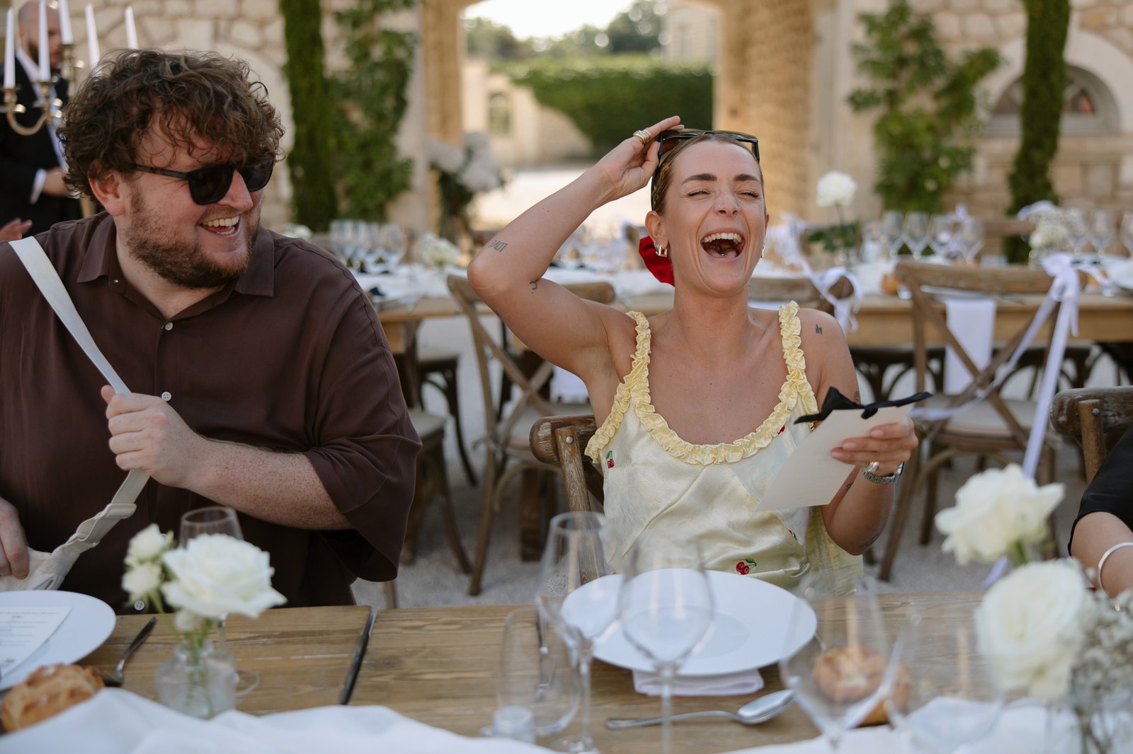 Two people sit at an outdoor table set for a formal event, laughing and holding menus or cards, with white flowers and glasses on the table.