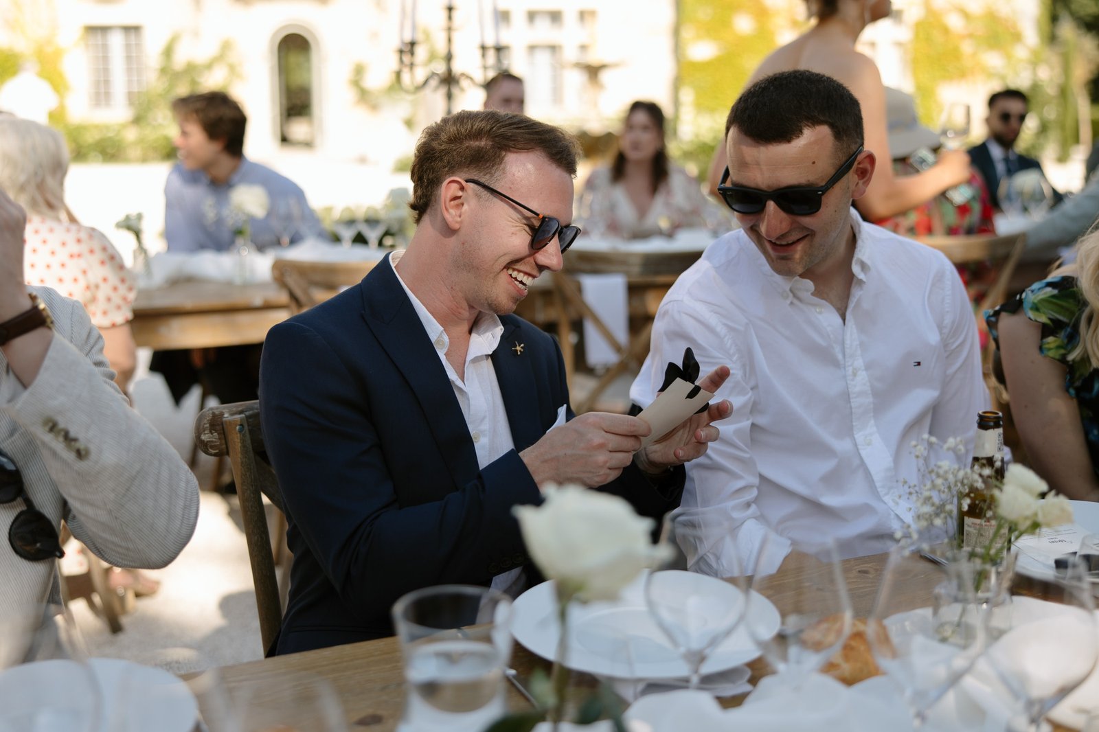 Two men in sunglasses sit at an outdoor table during a formal event, smiling and looking at a phone, with plates, glasses, and flowers on the table.