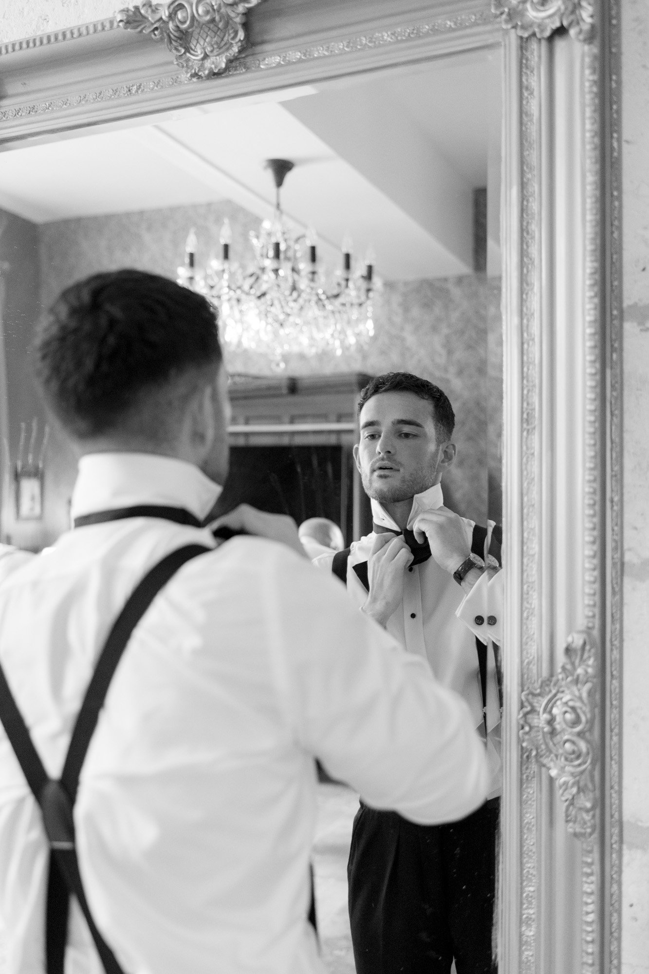 A man in formal attire stands in front of a large ornate mirror, adjusting his bow tie, with a chandelier reflecting in the background.