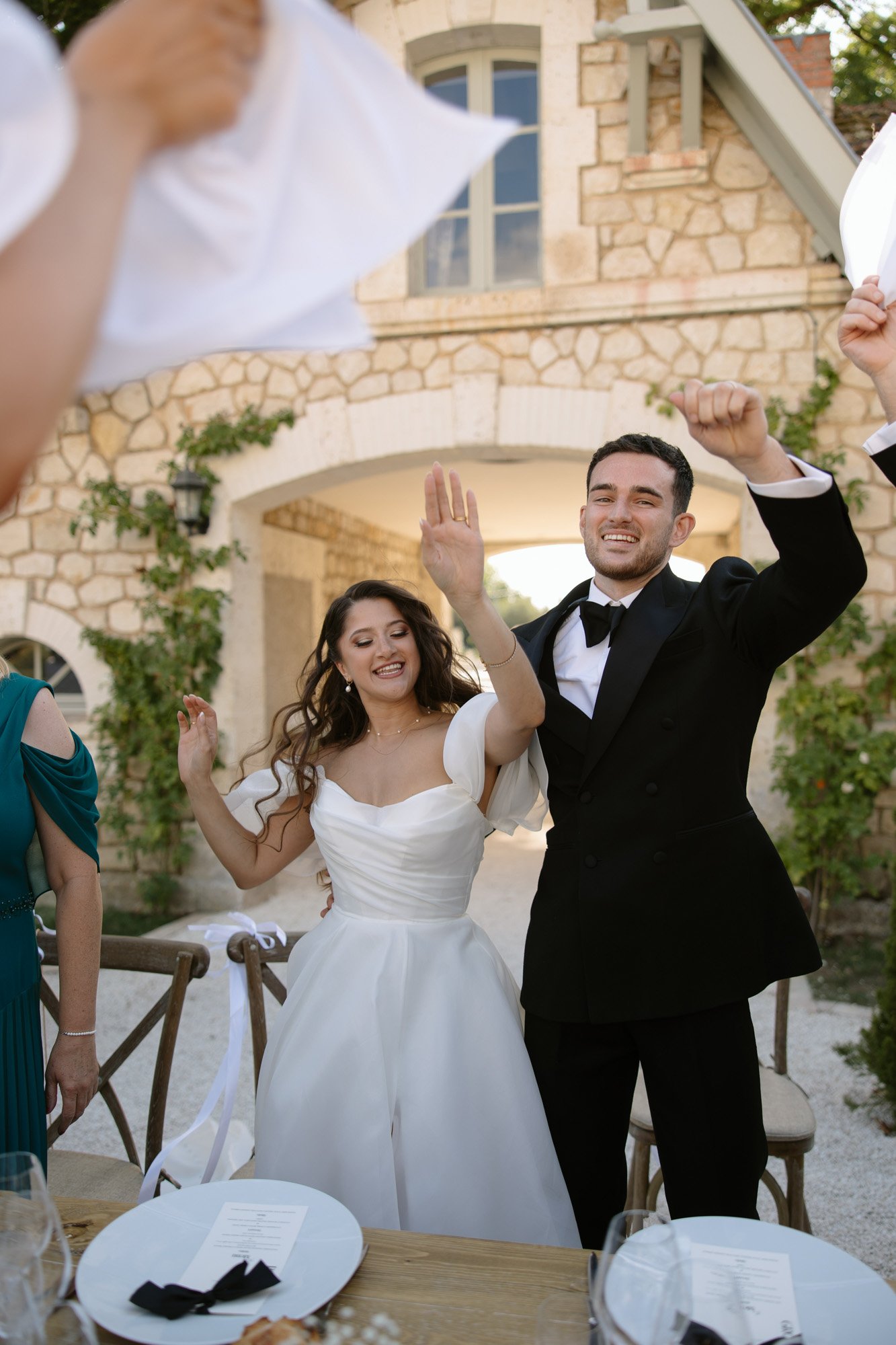 A bride and groom in formal attire stand and smile with raised arms at an outdoor wedding reception, with a stone building in the background. Chateau de la Couronne wedding.