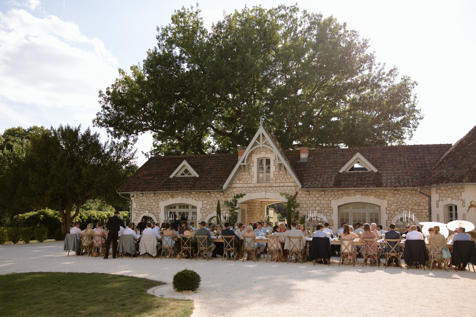 A group of people seated at long tables outdoors in front of a rustic stone building, dining together under a large tree on a sunny day. Chateau de la Couronne wedding.