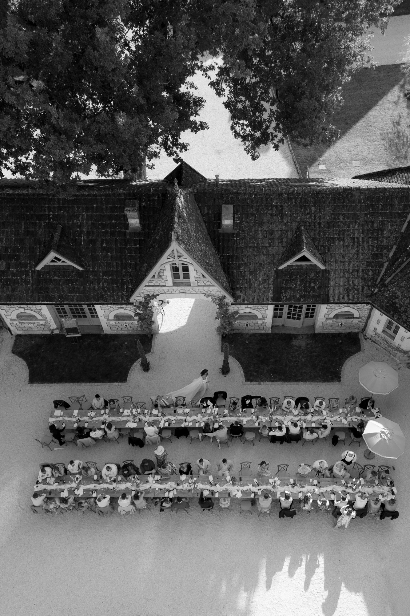 A large group of people sit at long banquet tables outdoors in front of a building, viewed from above in black and white.