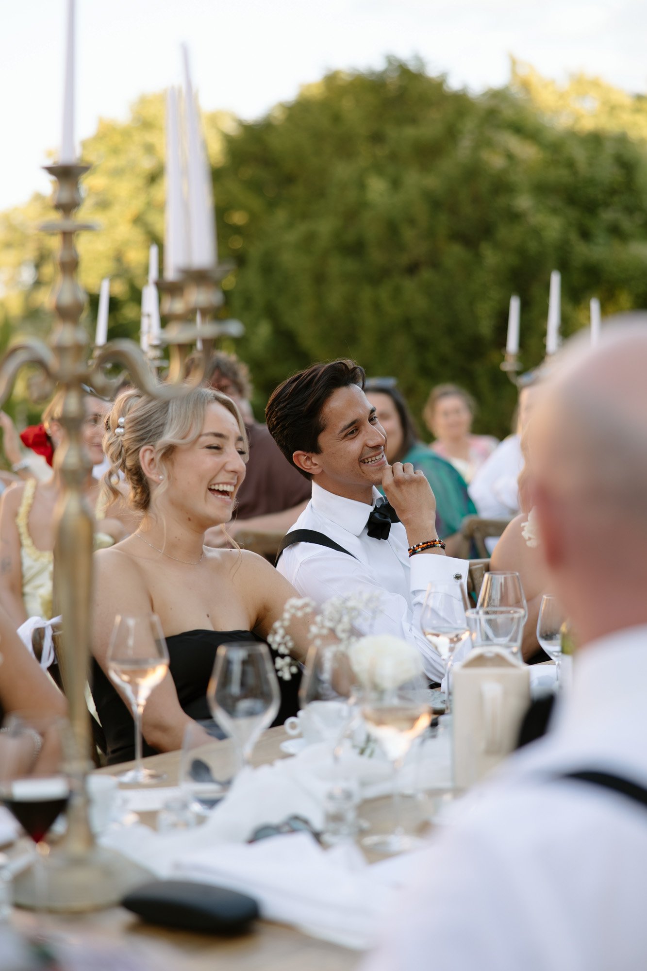 Two people dressed formally sit at an outdoor table, smiling and laughing among other guests during a daytime event. Candles, flowers, and glasses decorate the table. Chateau de la Couronne wedding.