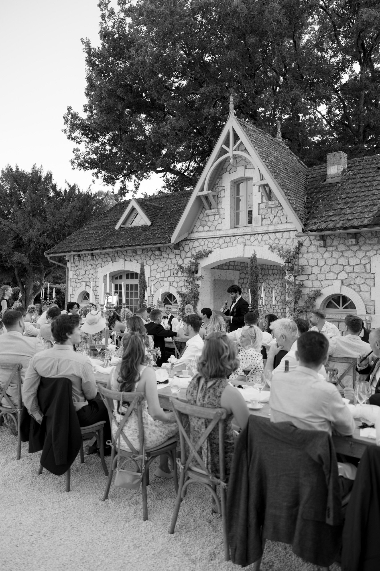 A group of people are seated at long tables outdoors in front of a stone house, listening to a person speaking at the head of the table. Chateau de la Couronne wedding.