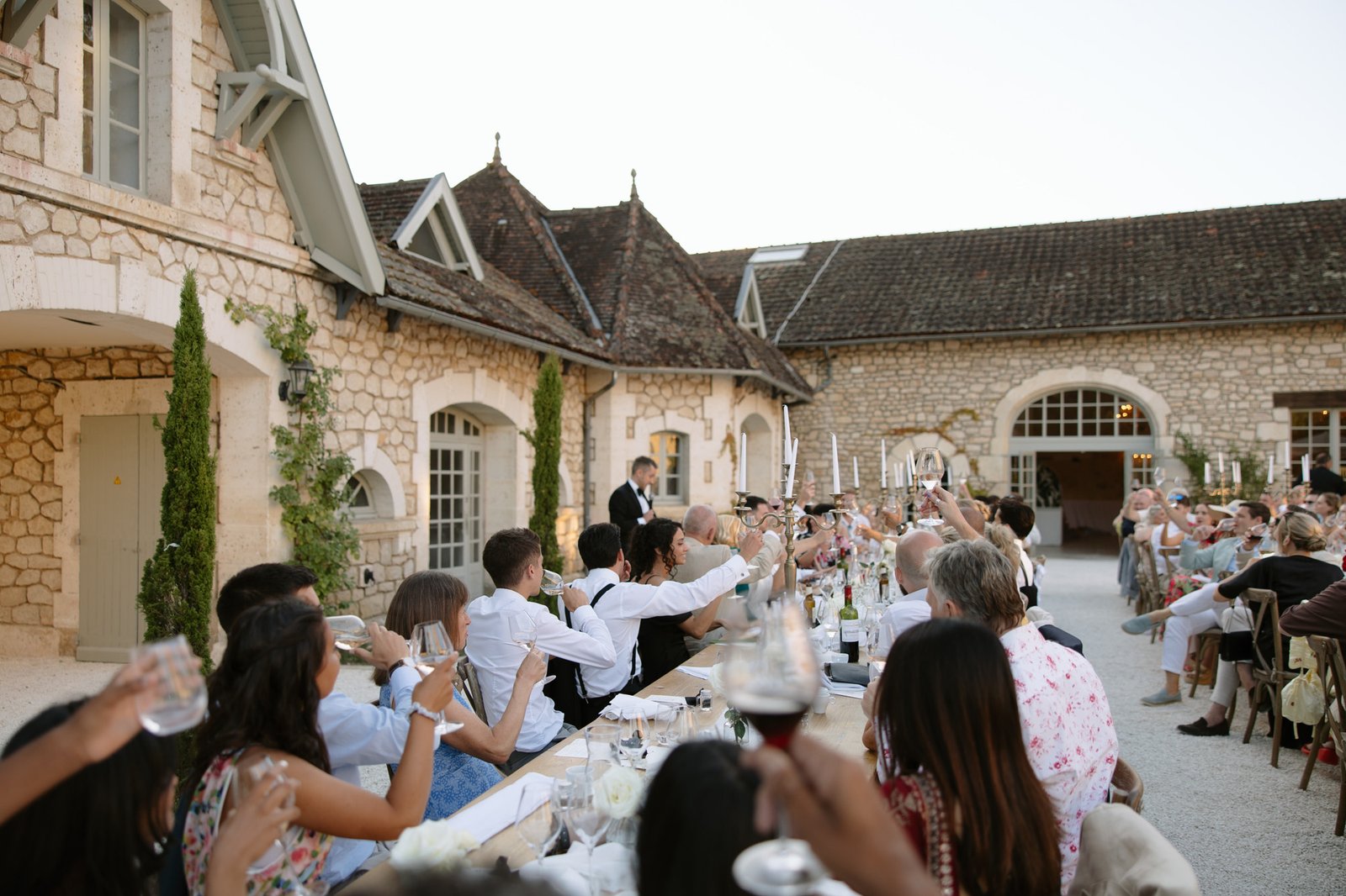 A group of people seated at long tables outdoors raise their glasses in a toast during an event at a rustic stone building.