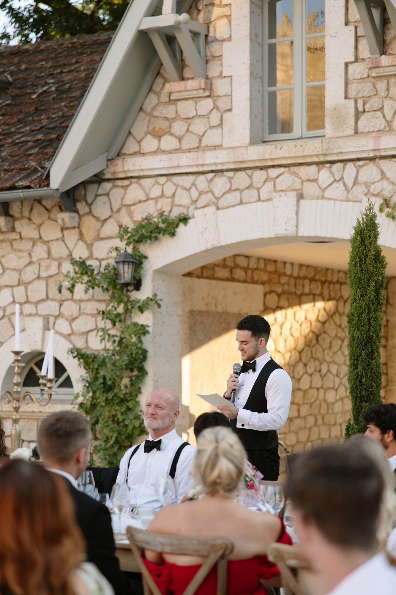 A man in formal attire stands and reads from a paper, addressing seated guests at an outdoor event in front of a stone building. Chateau de la Couronne wedding.
