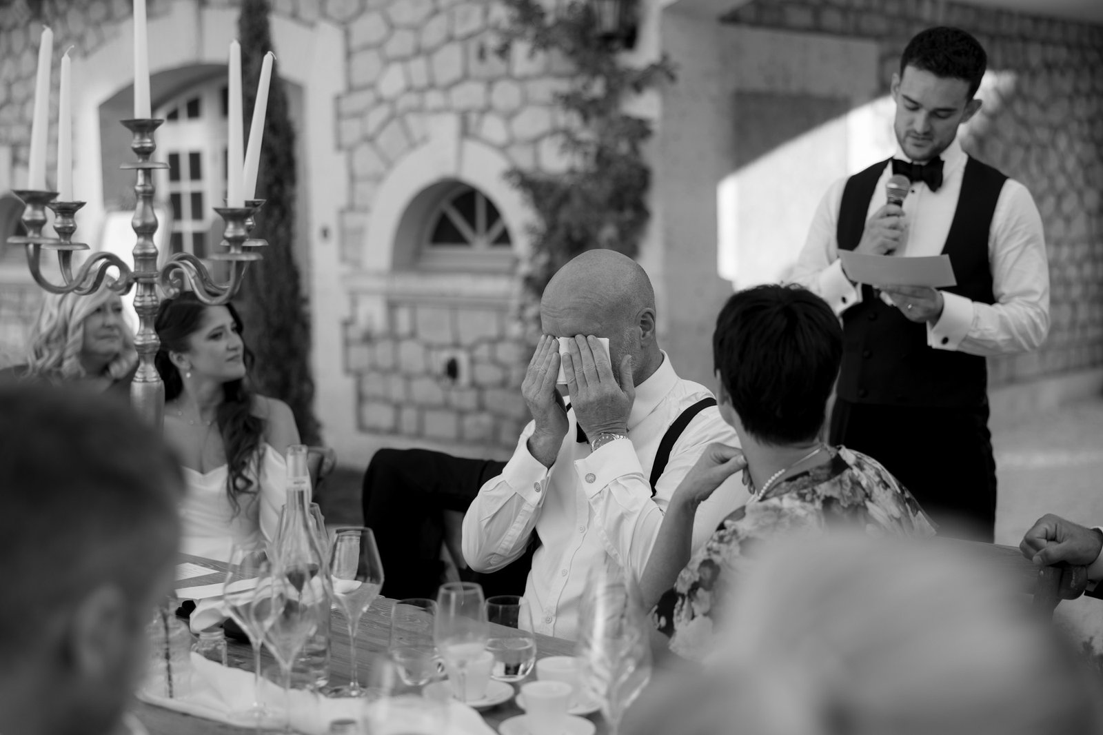 A man wipes his eyes with his hands at an outdoor wedding reception while a man in a vest and bow tie reads from a piece of paper. Guests sit around a decorated table.