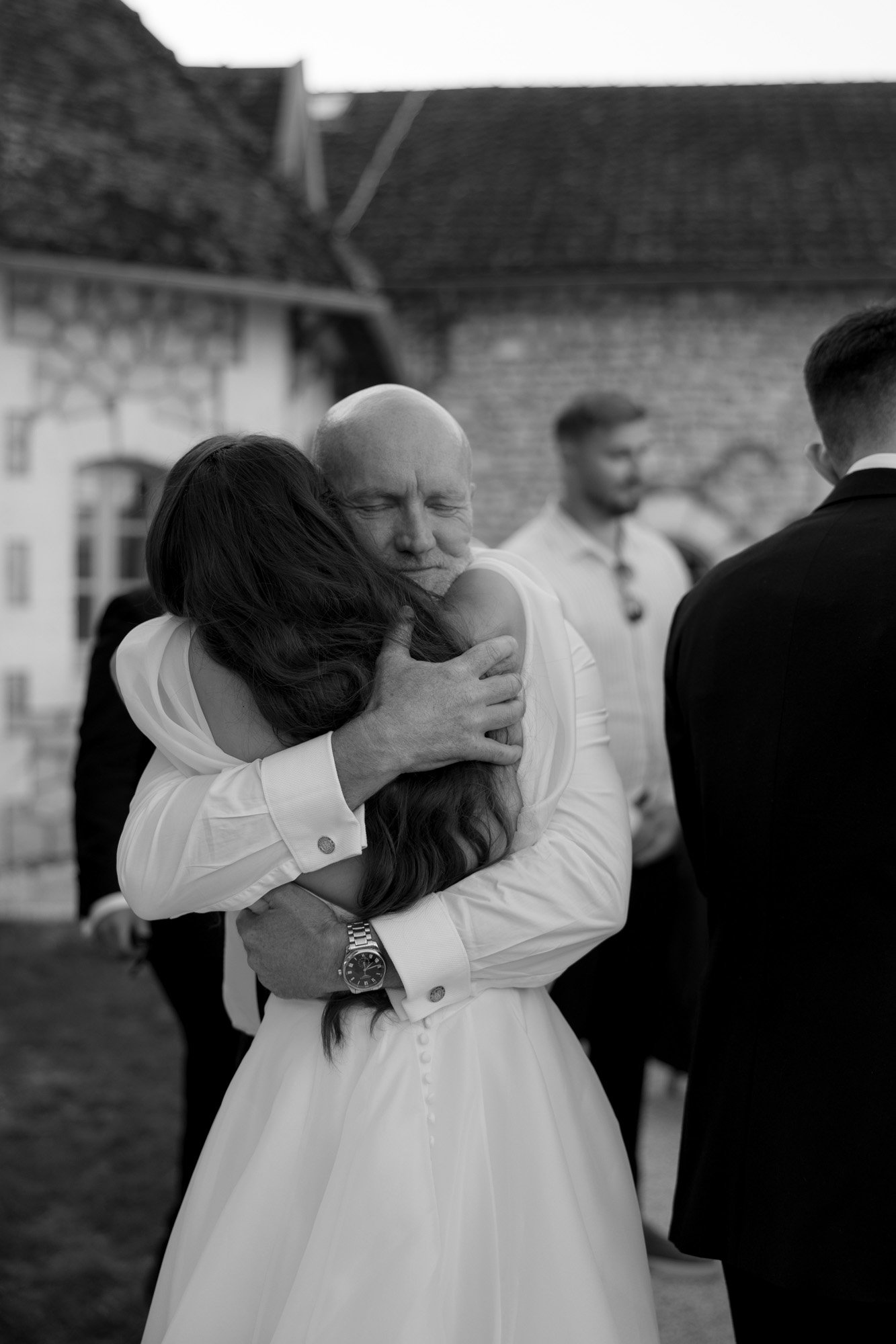 A man and a woman in formal attire embrace tightly outdoors; the woman wears a white dress, suggesting a wedding. The mans eyes are closed, and other people stand nearby. Chateau de la Couronne wedding.