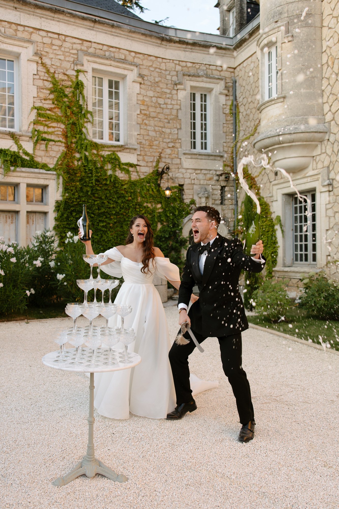 A bride and groom celebrate outside a stone building, enthusiastically opening champagne and spraying it next to a tower of empty glasses.