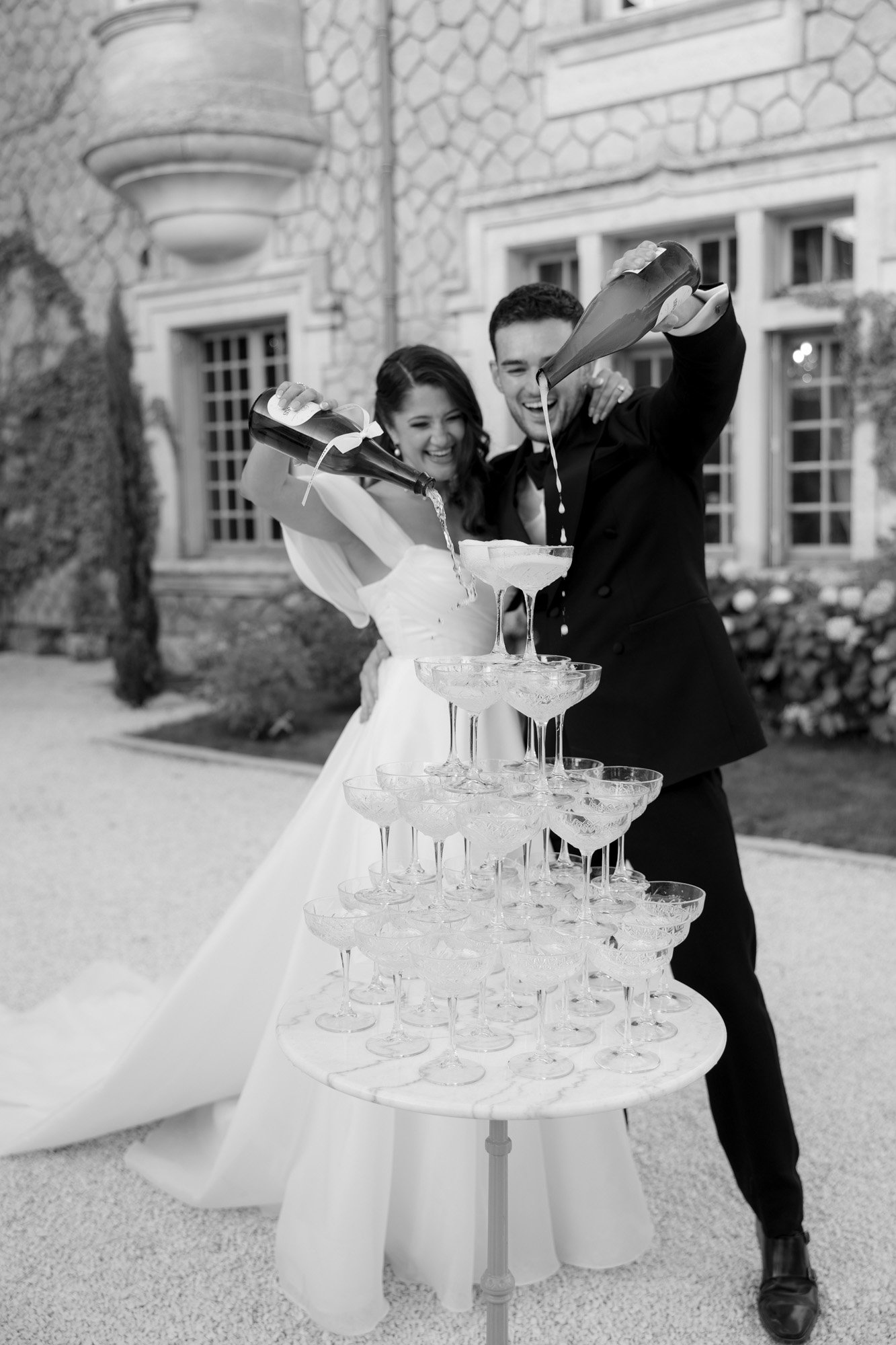 A bride and groom pour champagne into a tower of glasses outdoors in front of a stone building.