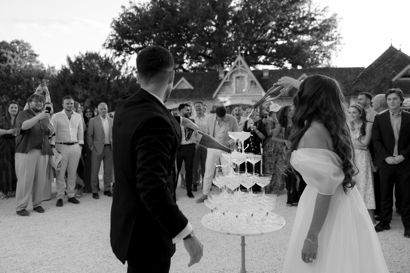 A bride and groom pour champagne into a tall pyramid of glasses outdoors as guests watch and take photos.