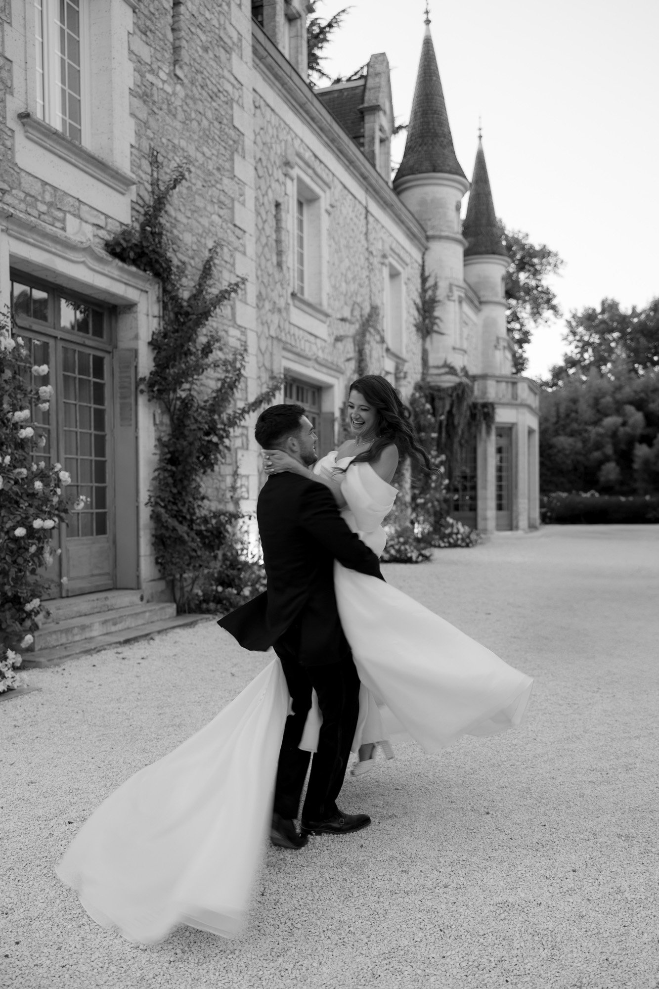 A couple in formal attire embrace and twirl outside a stone building with towers, surrounded by gardens.