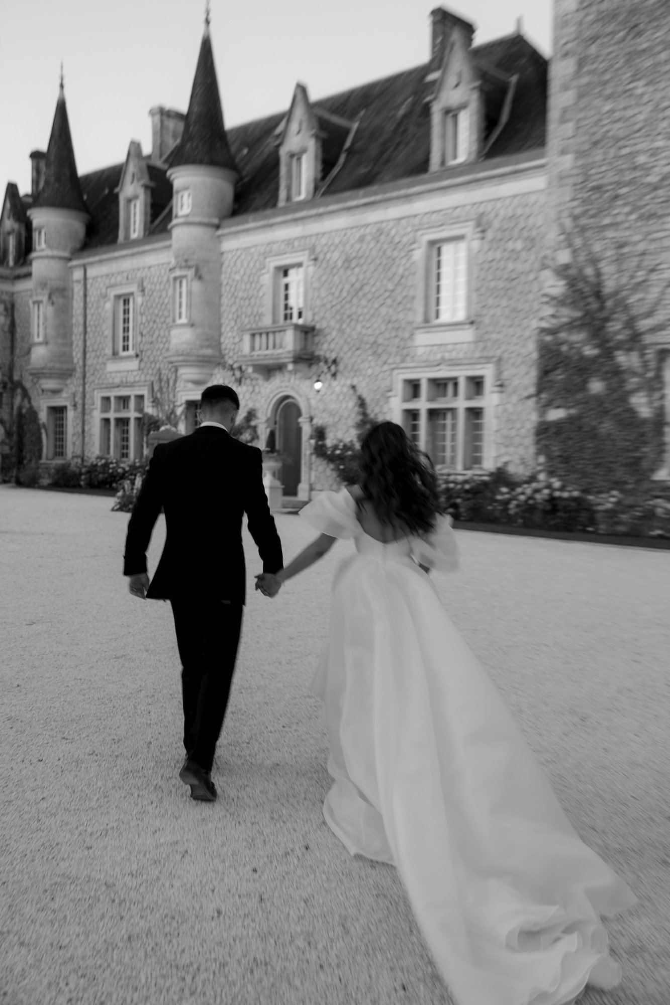 A bride and groom walk hand in hand toward a large stone building with turrets, possibly a castle, in an outdoor setting.