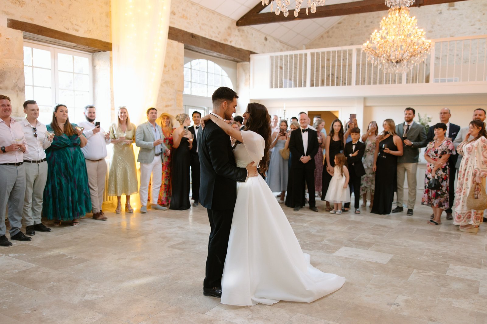 A bride and groom share their first dance in the center of a spacious, well-lit venue as guests stand around them watching.