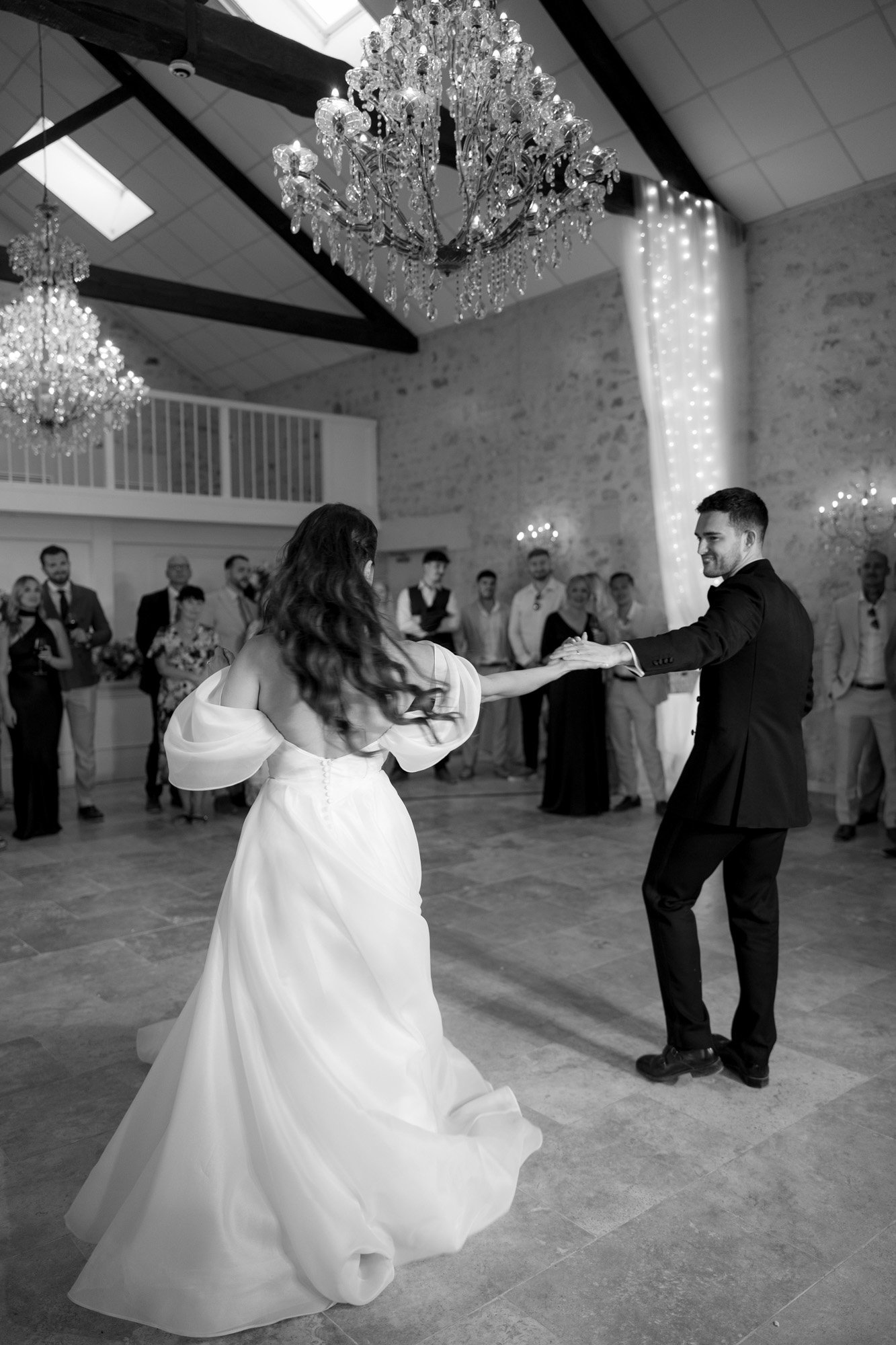 A bride and groom dance together in a spacious, elegant room with chandeliers, watched by guests in formal attire.