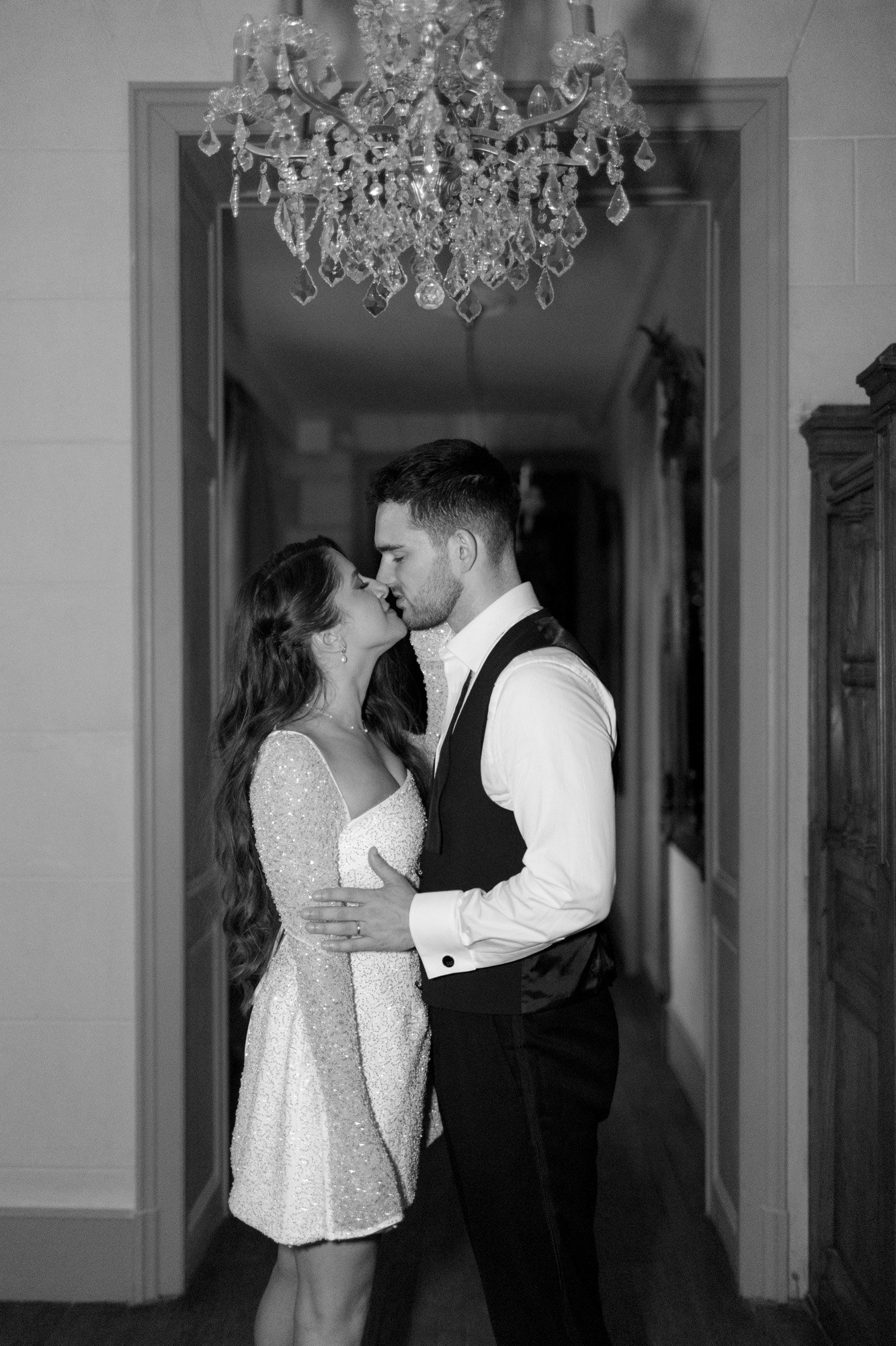 A couple dressed formally stands closely together in a hallway, gazing into each others eyes beneath a chandelier.
