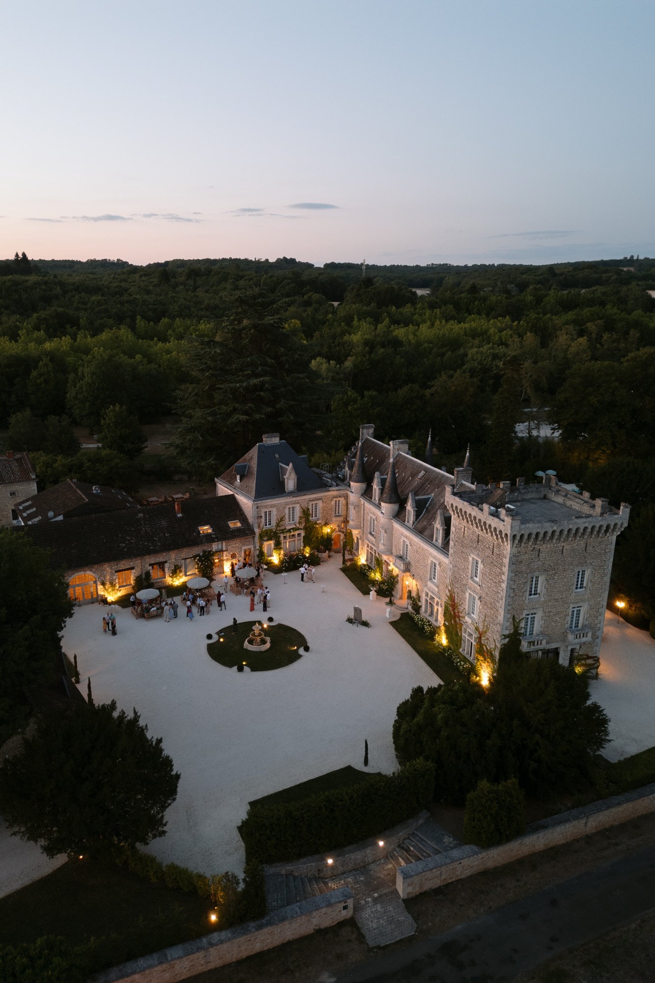 Aerial view of a stone château with lit windows and courtyard, hosting an evening event; surrounded by trees and greenery at dusk.