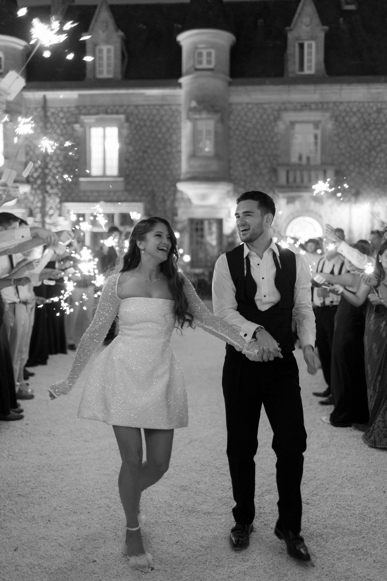 A couple walks hand-in-hand through a crowd holding sparklers at night, in front of a large stone building, both smiling and dressed in formal attire.