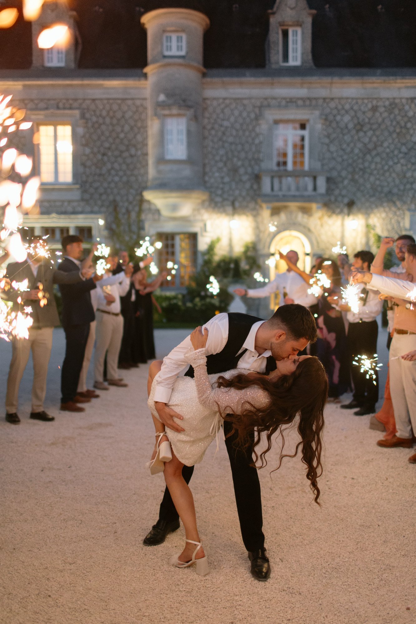 A couple shares a kiss during a dip as guests holding sparklers celebrate outside an elegant stone building in the evening.