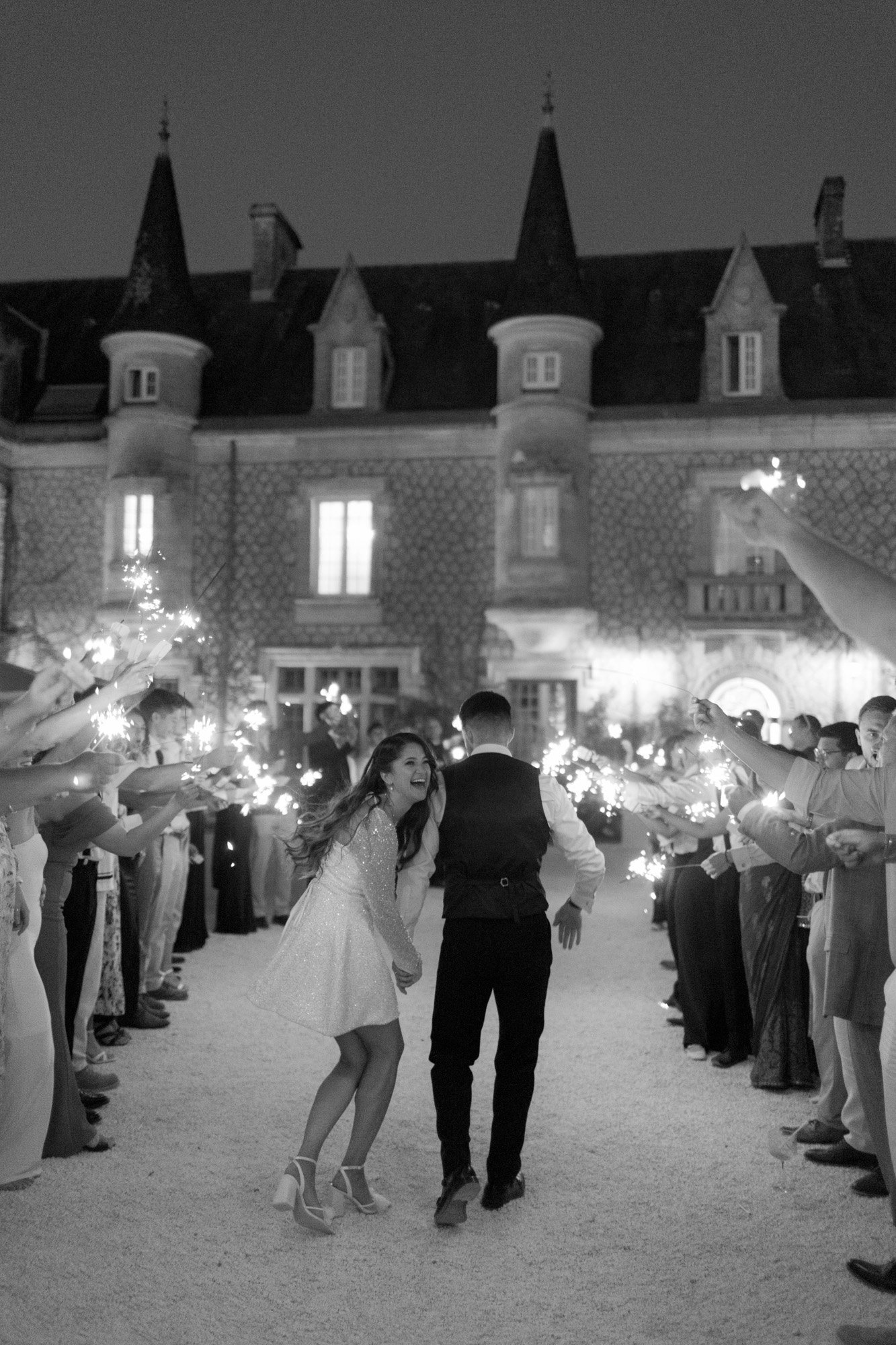 A bride and groom walk through a crowd holding sparklers outside a large stone building at night.