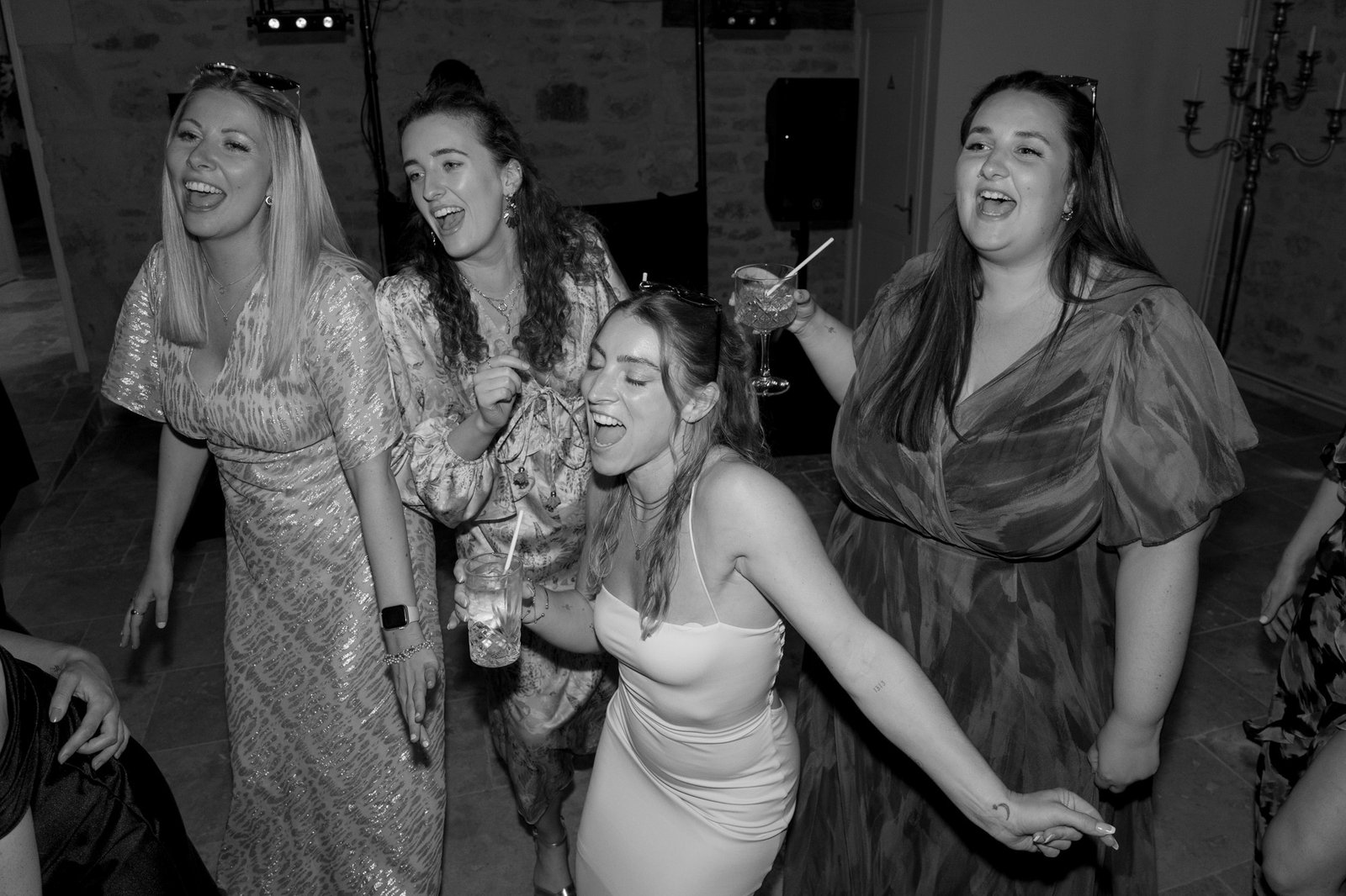 Four women dressed in formal attire sing and laugh together at an indoor event, each holding a drink.
