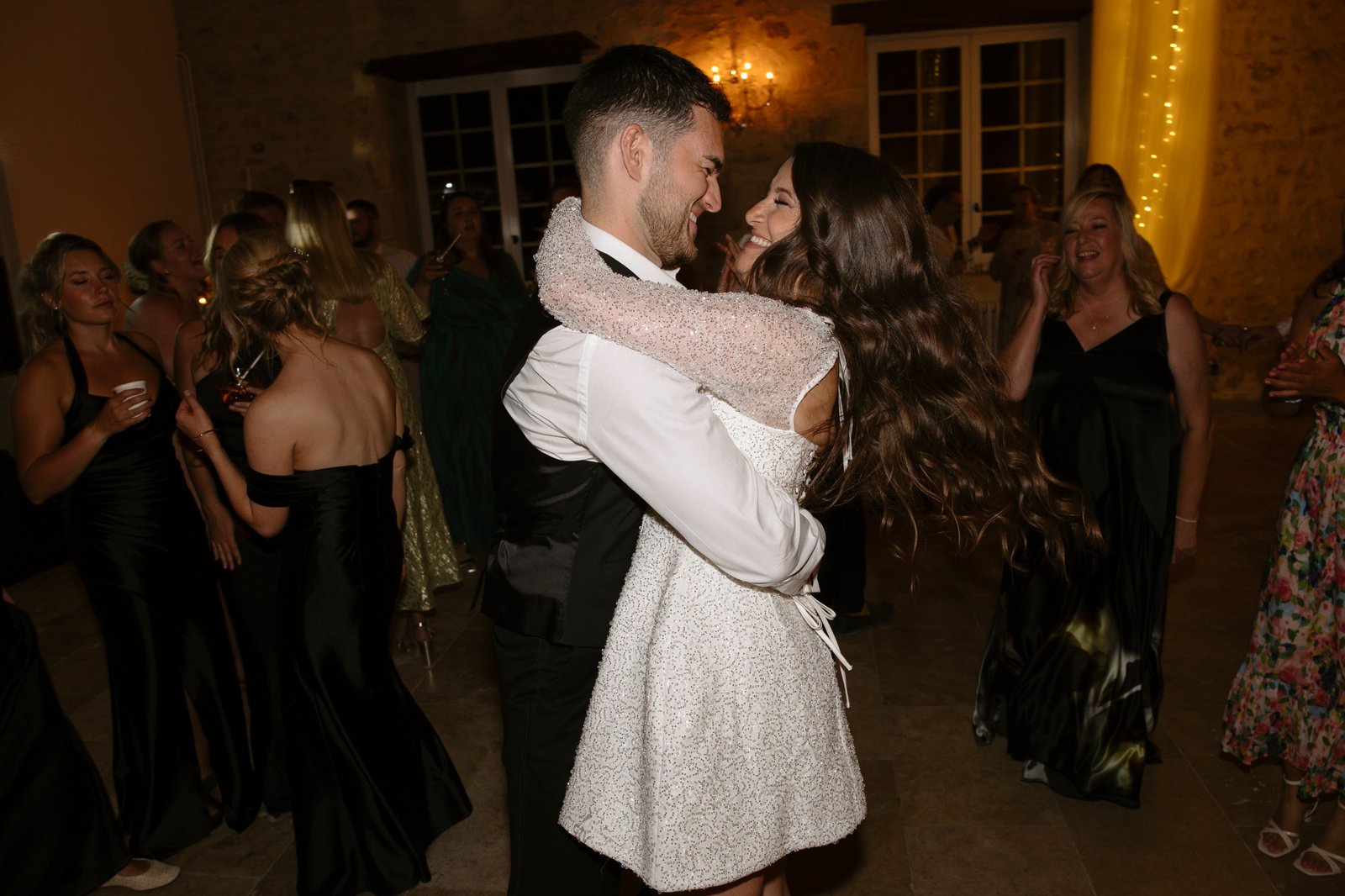 A couple embraces and smiles while dancing at an indoor event, surrounded by guests dressed in formal attire.