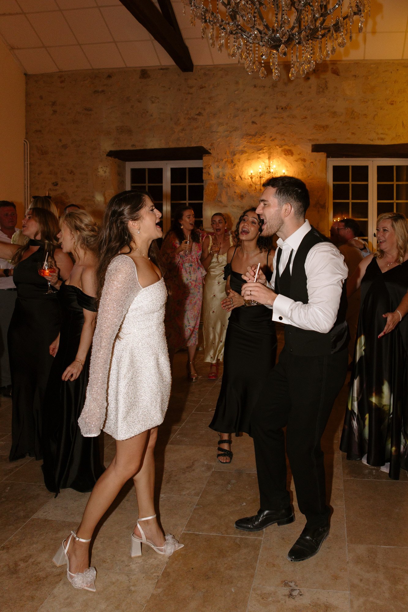 A group of people dressed in formal attire dance and sing together in a warmly lit room with stone walls and a chandelier.