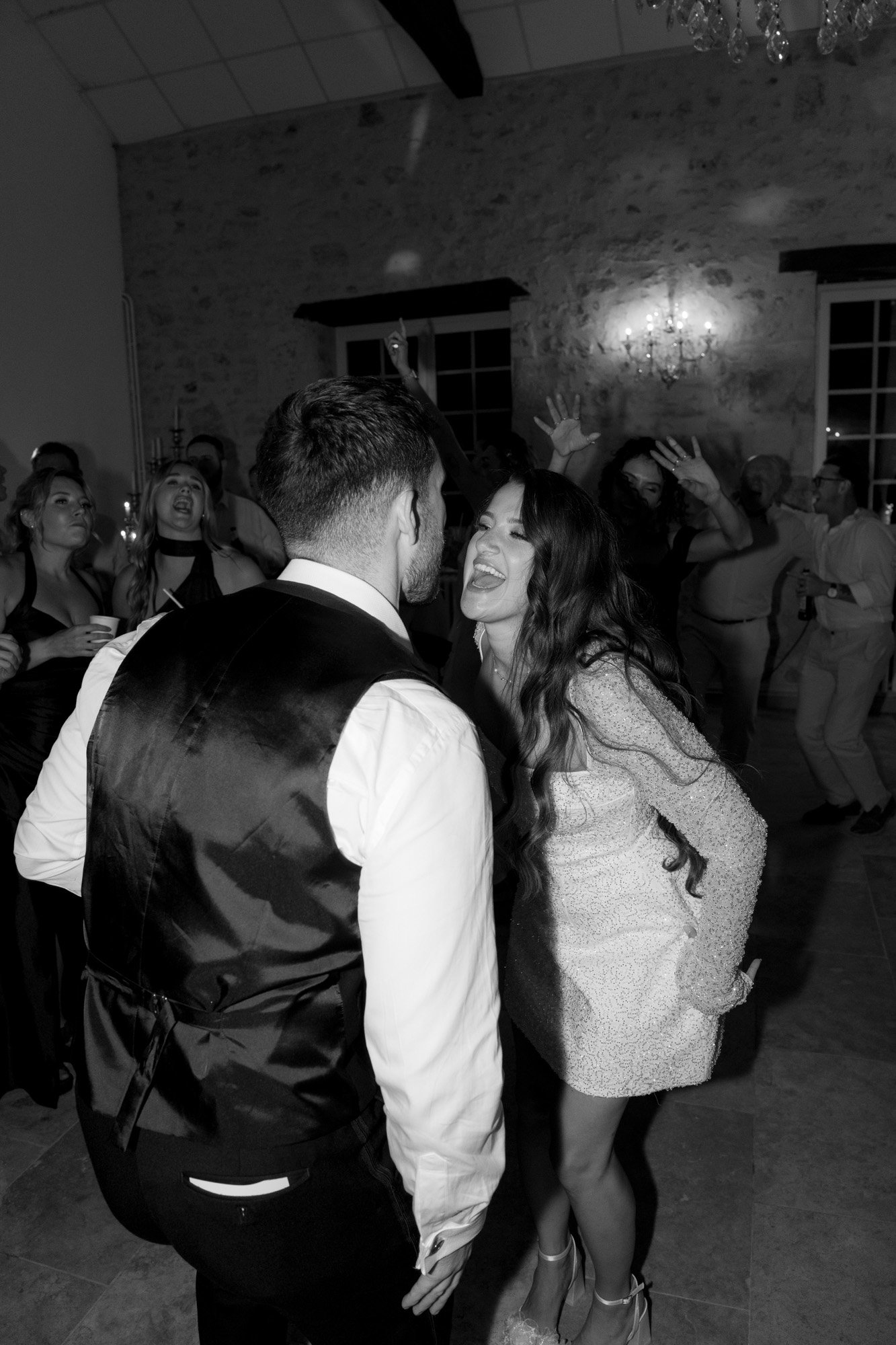 A woman in a dress joyfully sticks out her tongue at a man in a vest during a lively indoor party with people dancing in the background.
