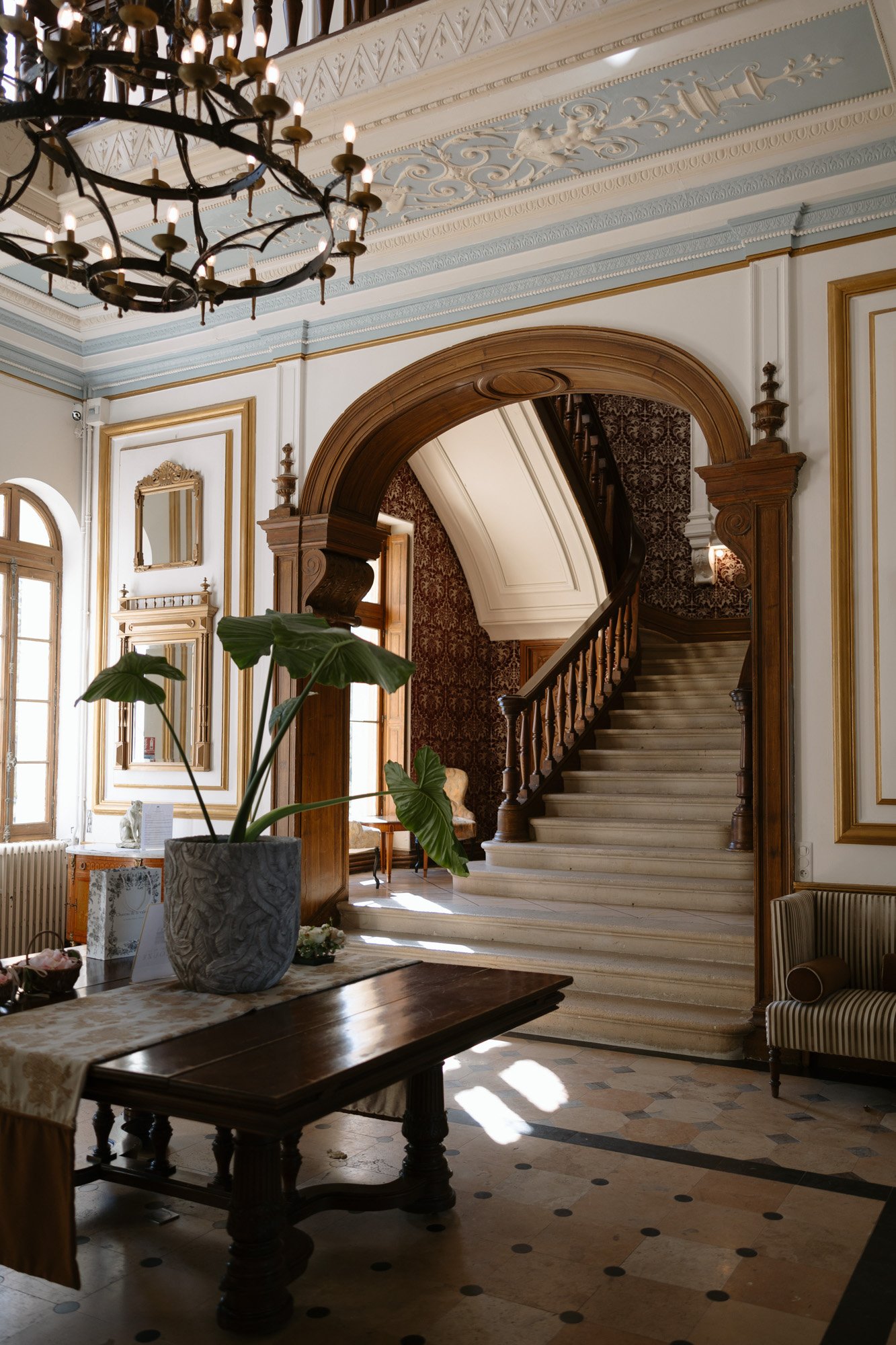 A grand foyer with a wooden staircase, ornate archway, chandelier, large plant in a pot on a dark wood table, and natural light from tall windows.