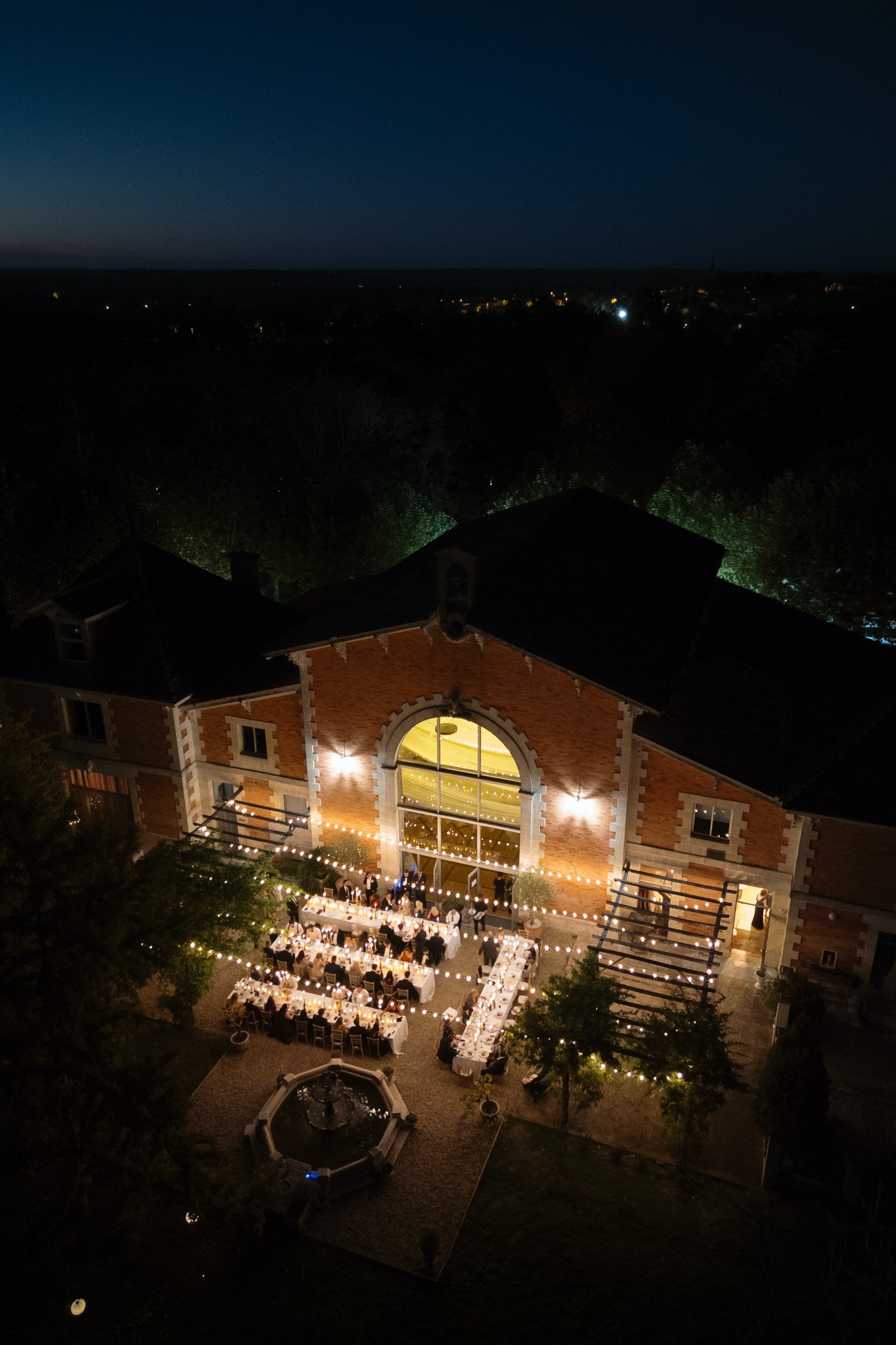 Aerial view of an outdoor evening event with long dining tables set up in front of a large building illuminated by lights. Chateau de la Valouze wedding.